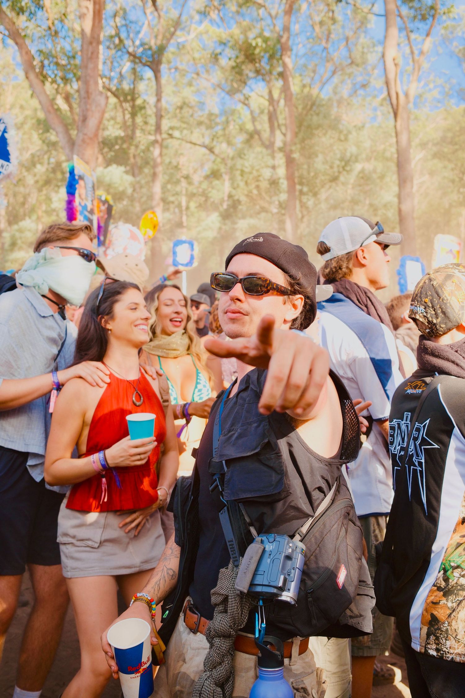 Man in black cap and sunglasses pointing at camera surrounded by festival crowd outdoors.