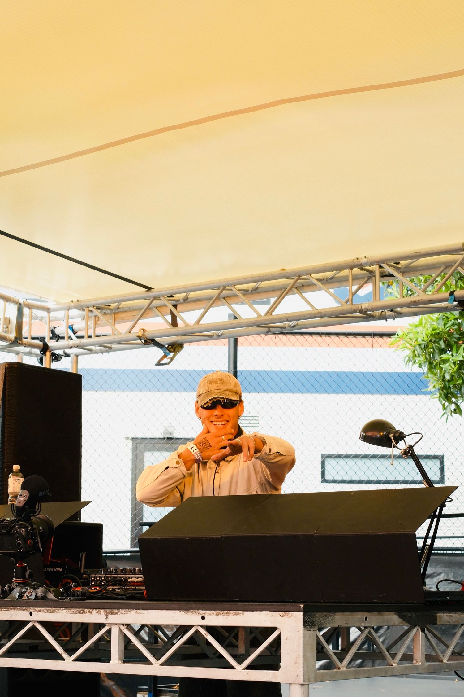 DJ in cap and sunglasses performing at an outdoor festival stage under a canopy tent.