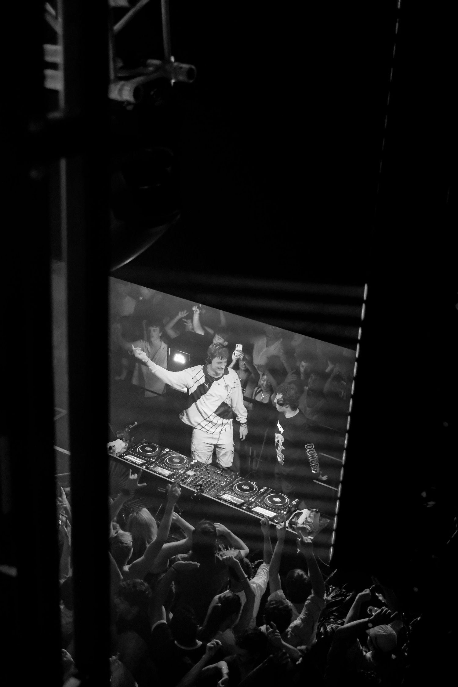 Black and white photo of a DJ performing on stage with crowd cheering below at a nightclub event.