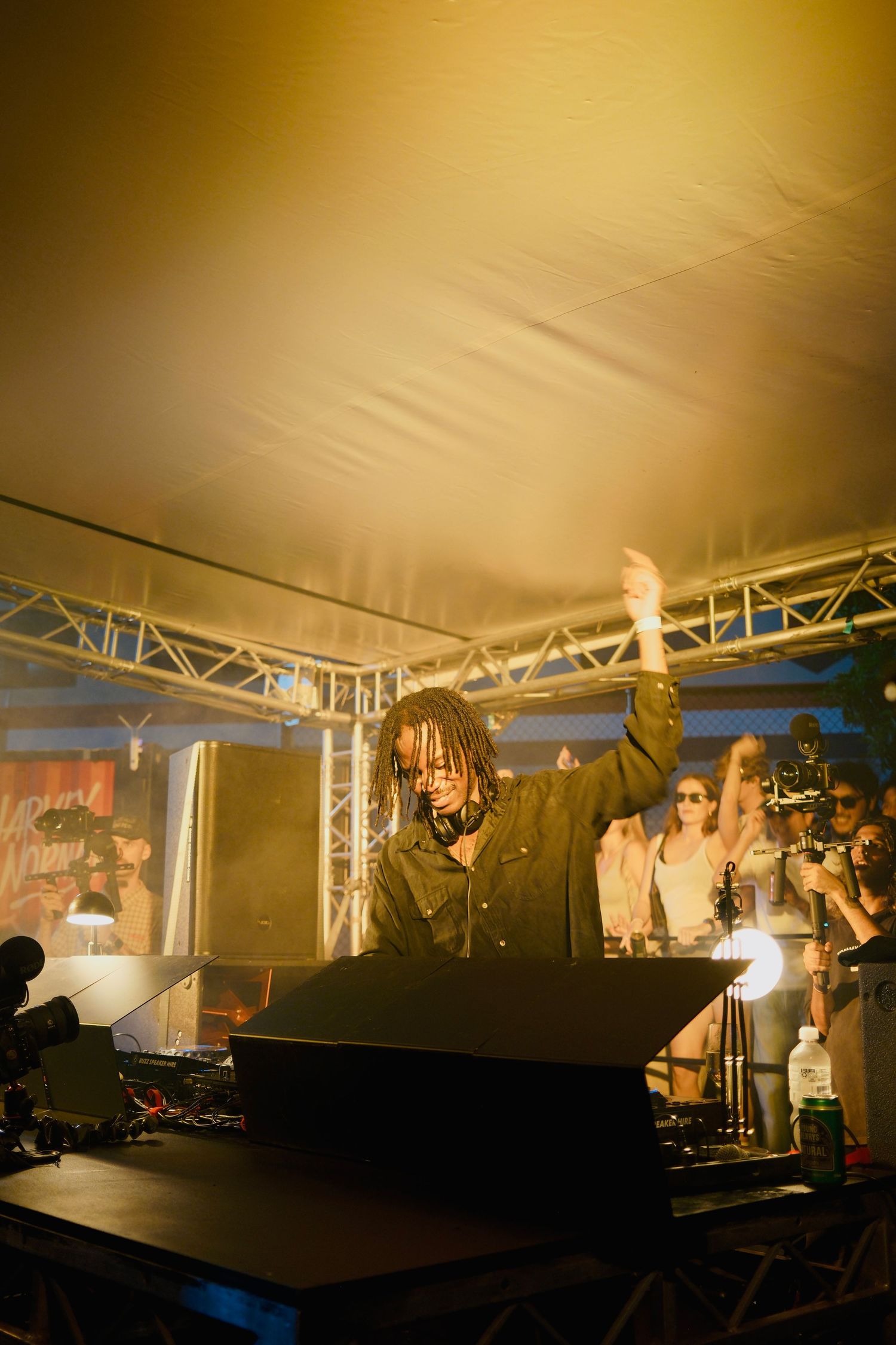 DJ with dreadlocks raising hand at outdoor festival decks under golden tent lighting.