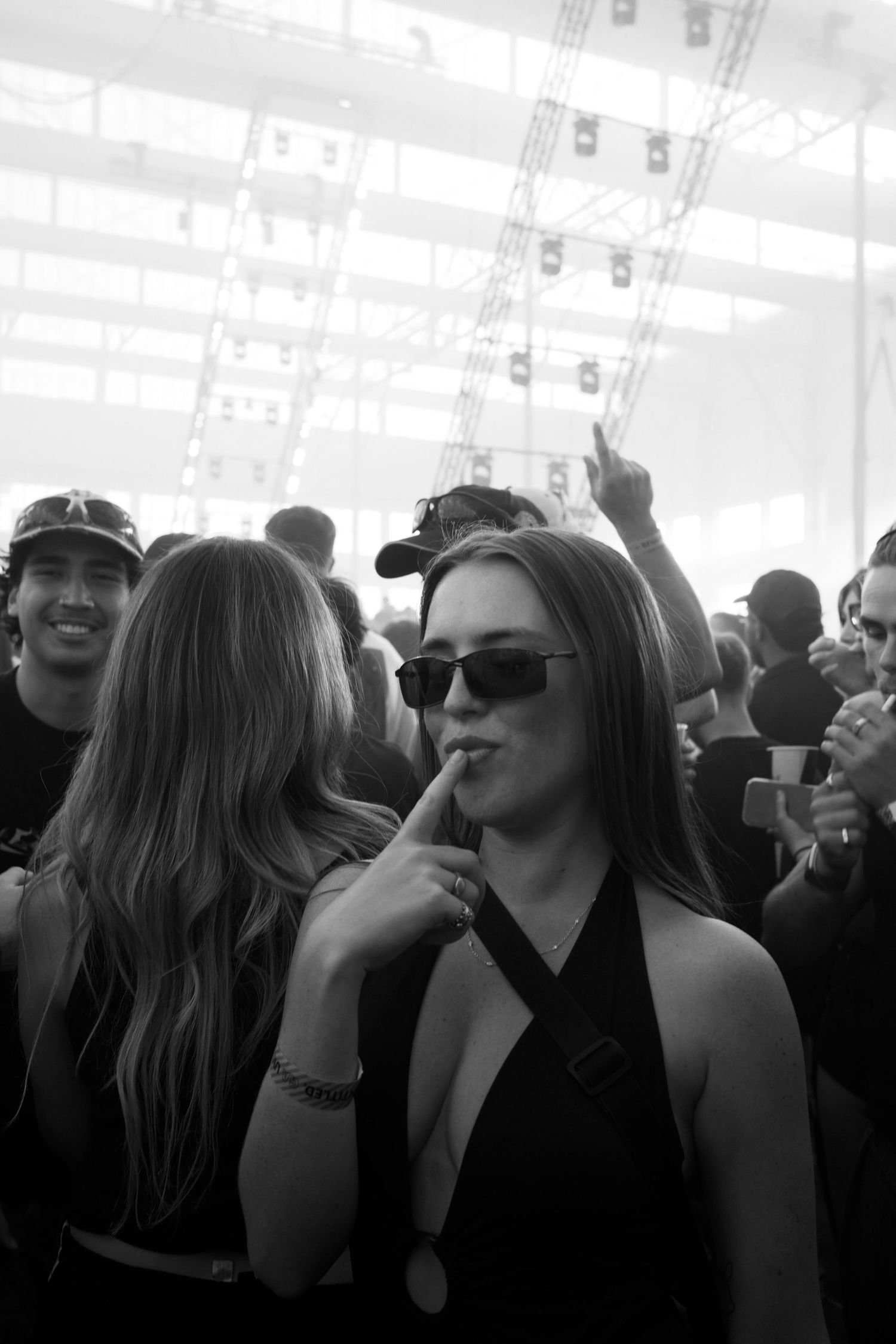 Black and white photo of a woman in sunglasses posing at a crowded indoor music festival.