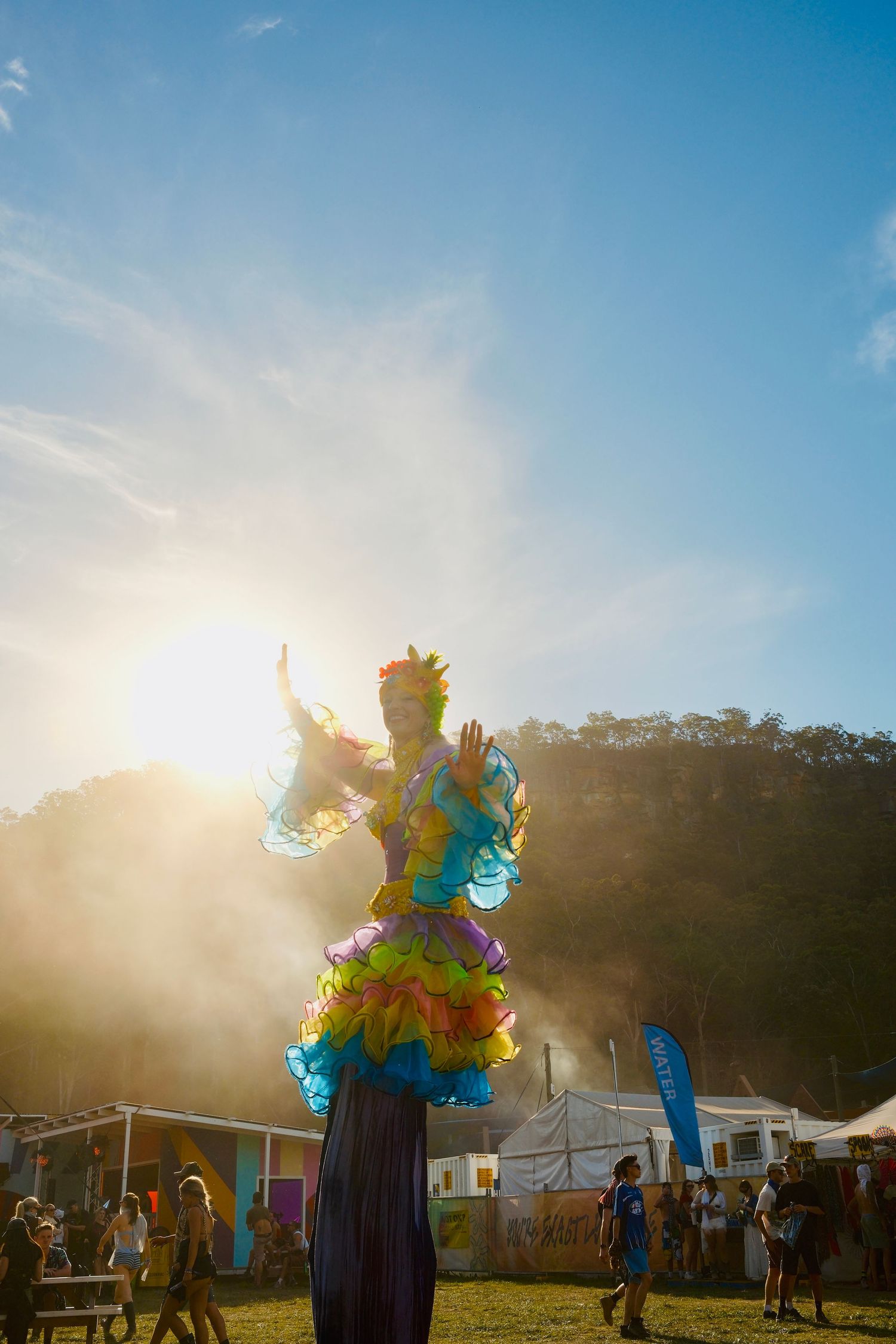 Colorful stilt performer in rainbow costume dancing at a sunny outdoor music festival.