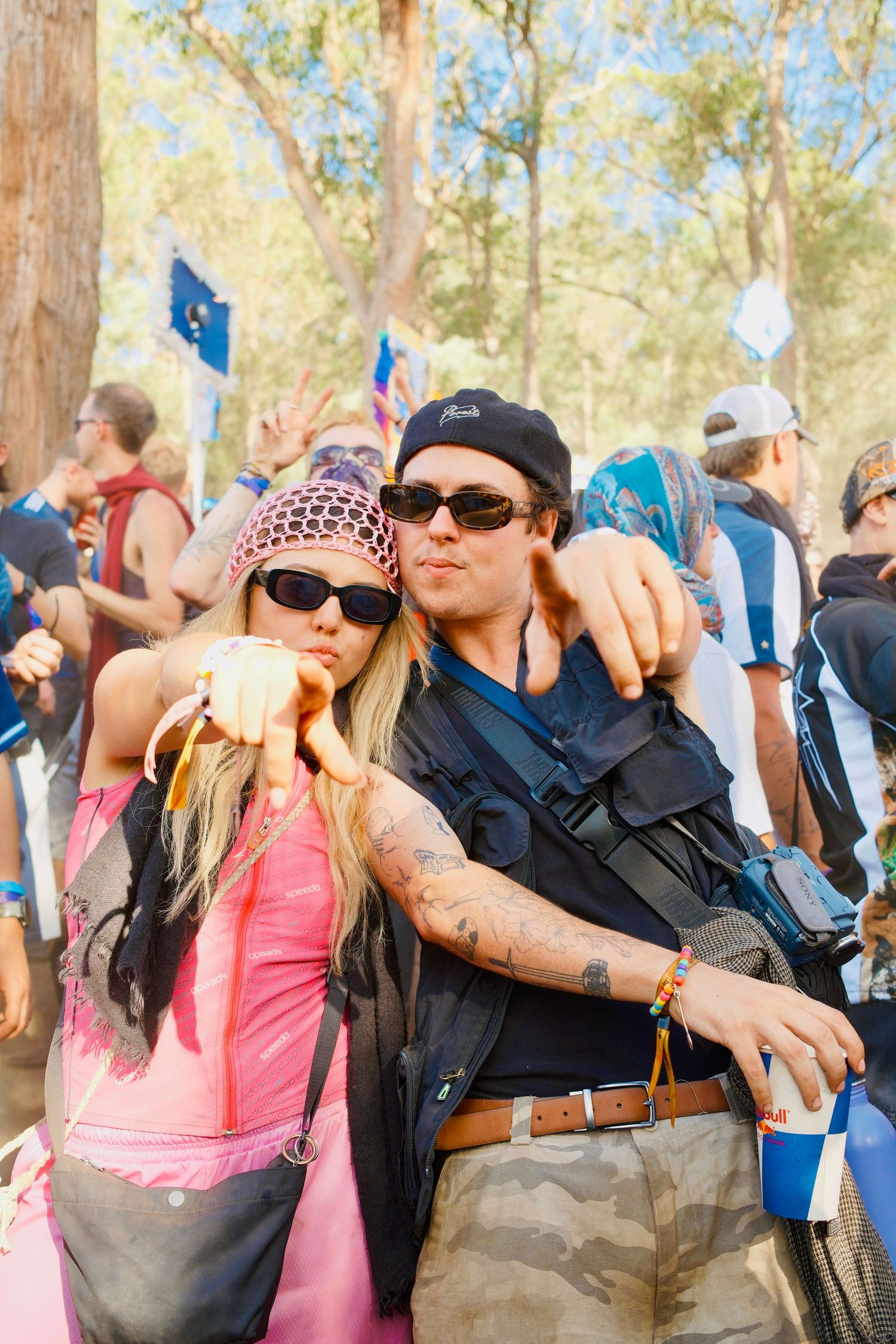 Couple in festival attire posing together at an outdoor music festival among eucalyptus trees.