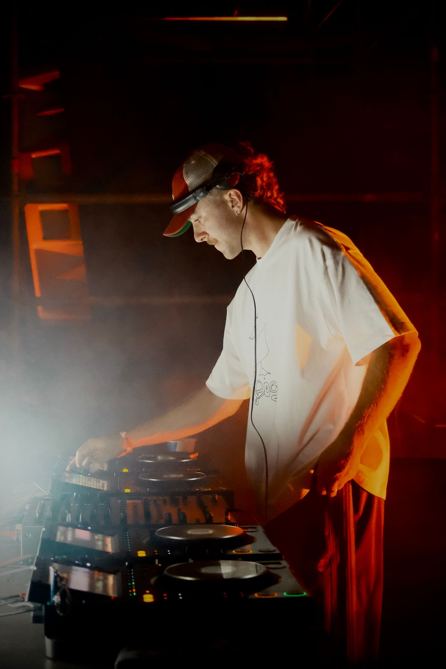 DJ in a white t-shirt and cap mixing tracks on turntables under red and orange stage lighting with smoke effects.