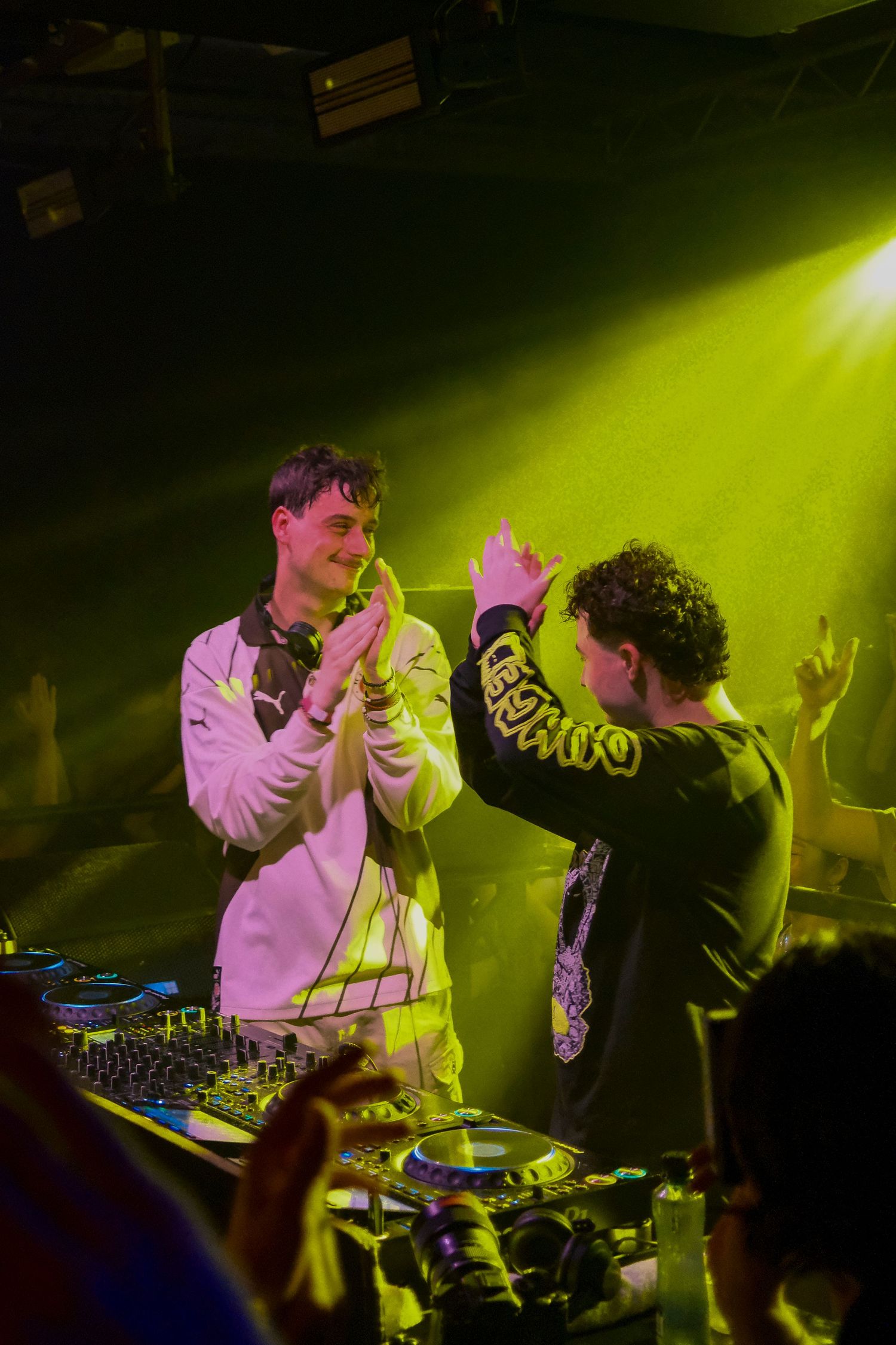 Two DJs high-fiving at a DJ booth under green stage lights at a lively nightclub event.