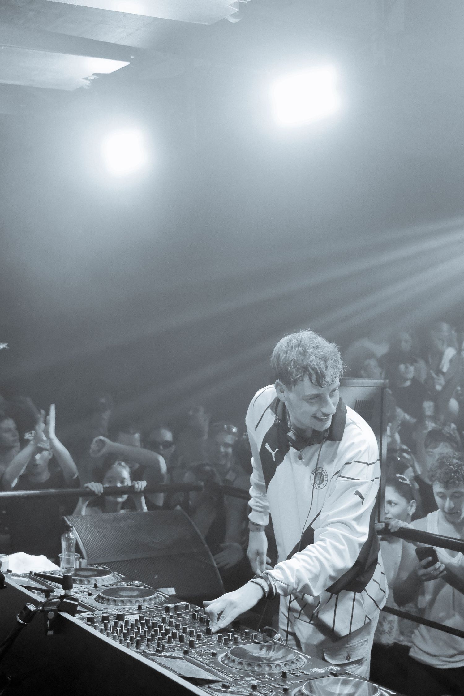 DJ performing at a nightclub with crowd behind, bright stage lights and mixing equipment in black and white photo.