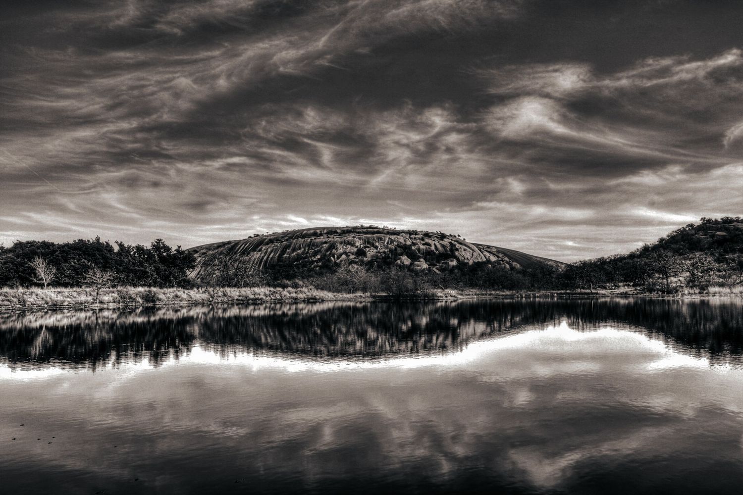 Black-and-white landscape photograph of a rocky hill reflected in still water, emphasizing texture, balance, and the photographer’s roots in
