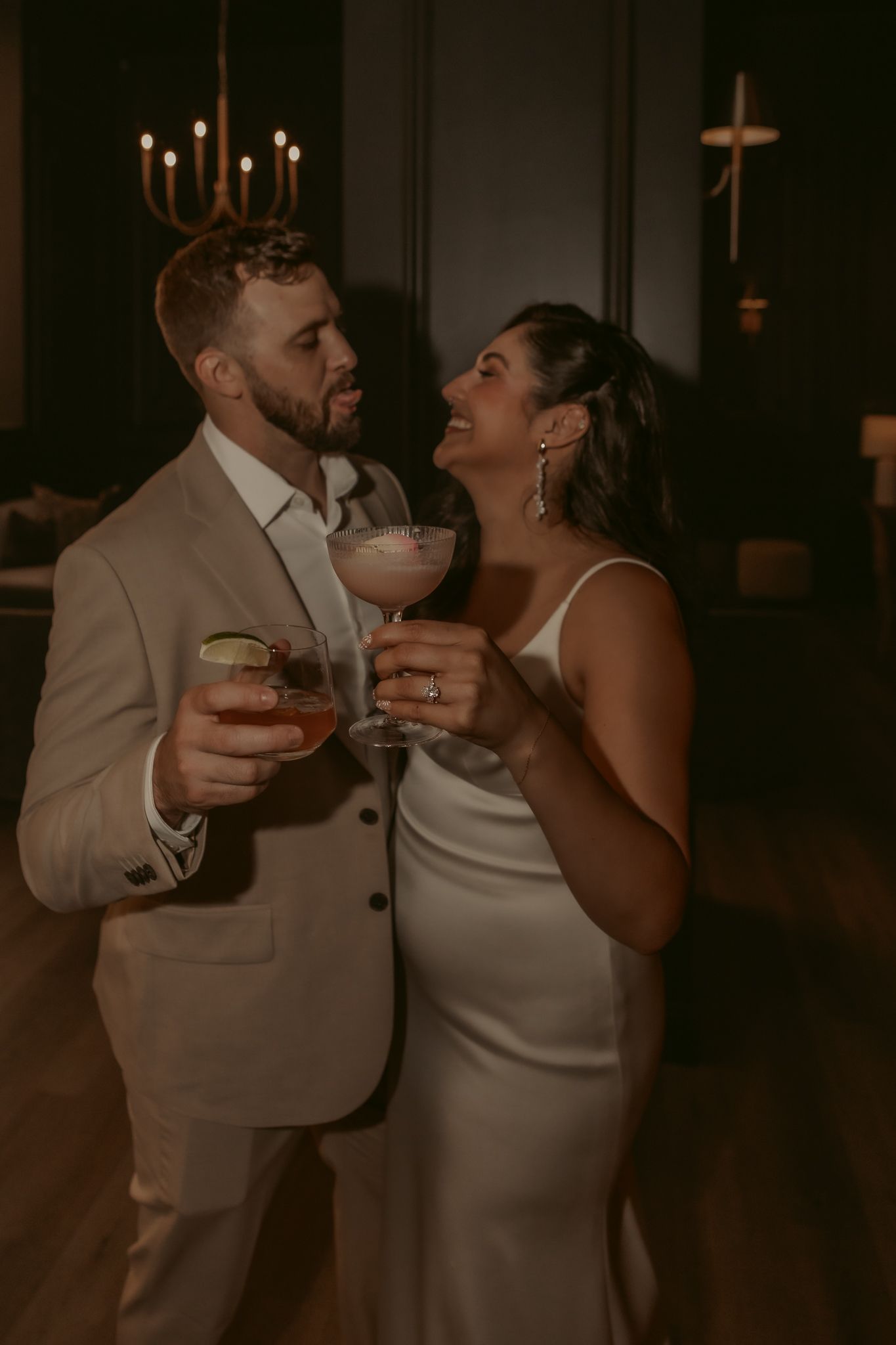 Smiling couple in a tan suit and white satin gown clink cocktail glasses in a warmly lit, candlelit room at an elegant evening celebration.