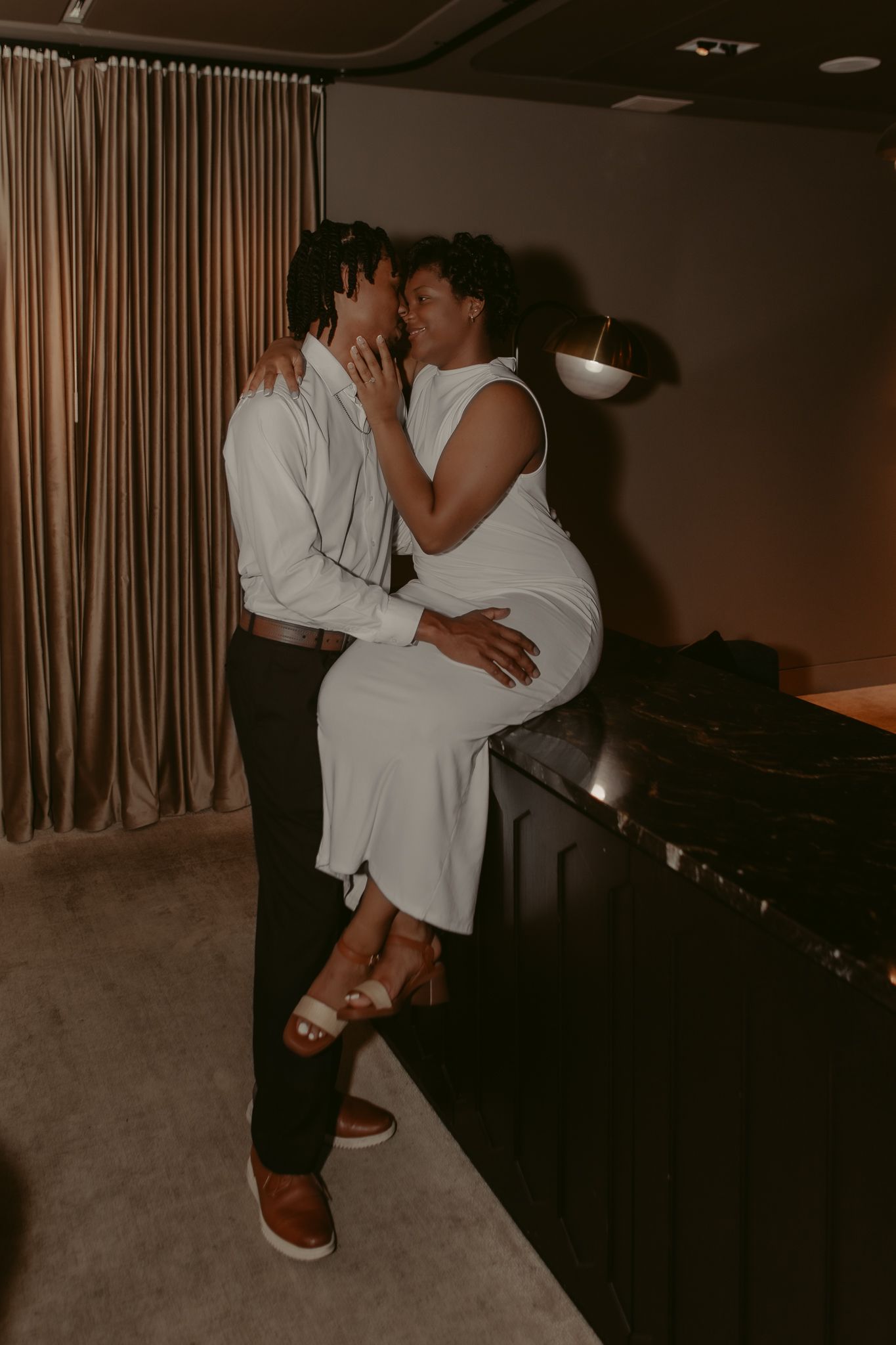 Couple in white outfits embracing and kissing beside a dark marble counter in a softly lit room with draped curtains.