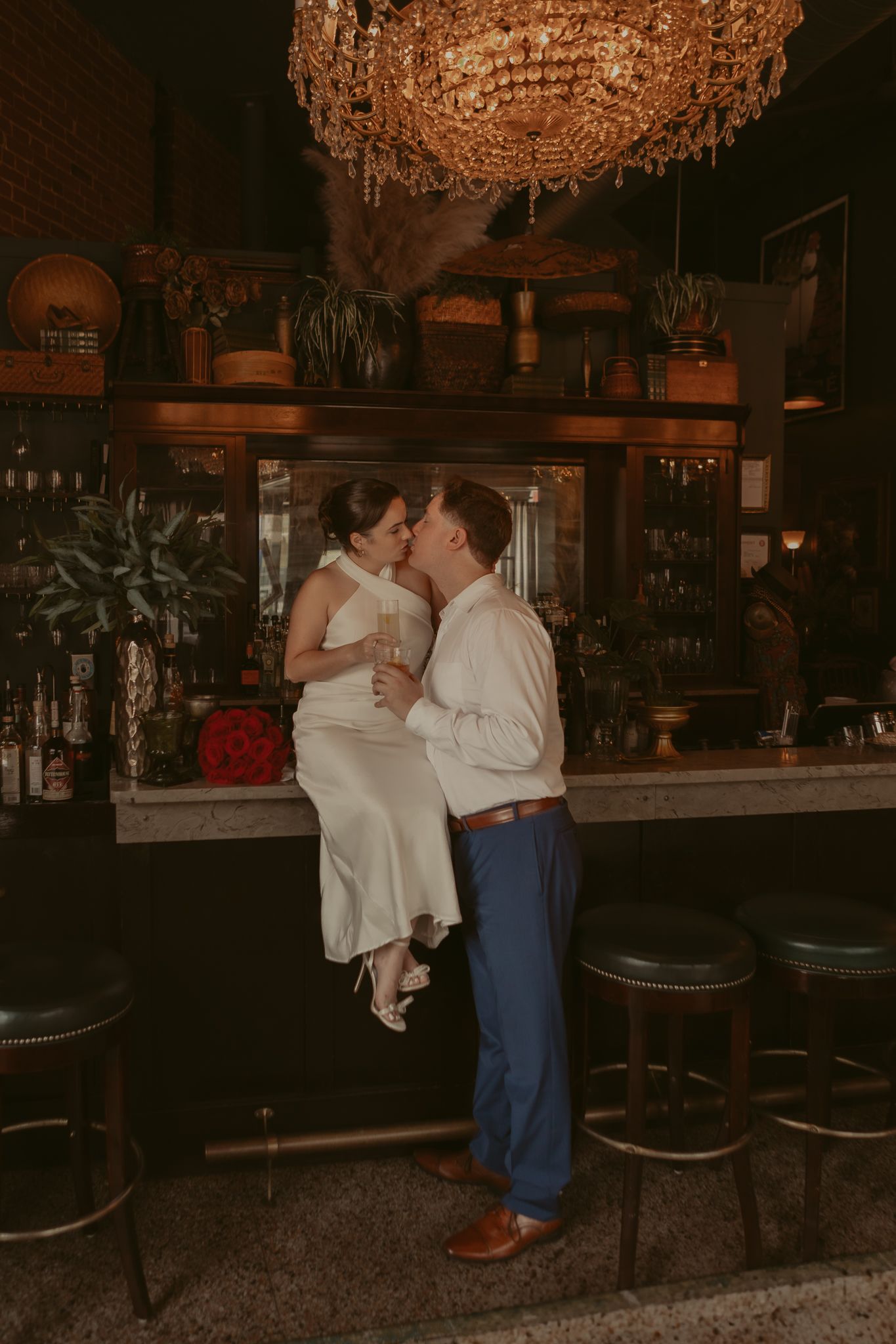 Bride sits on bar counter as groom kisses her in elegant dimly lit vintage bar with chandelier.
