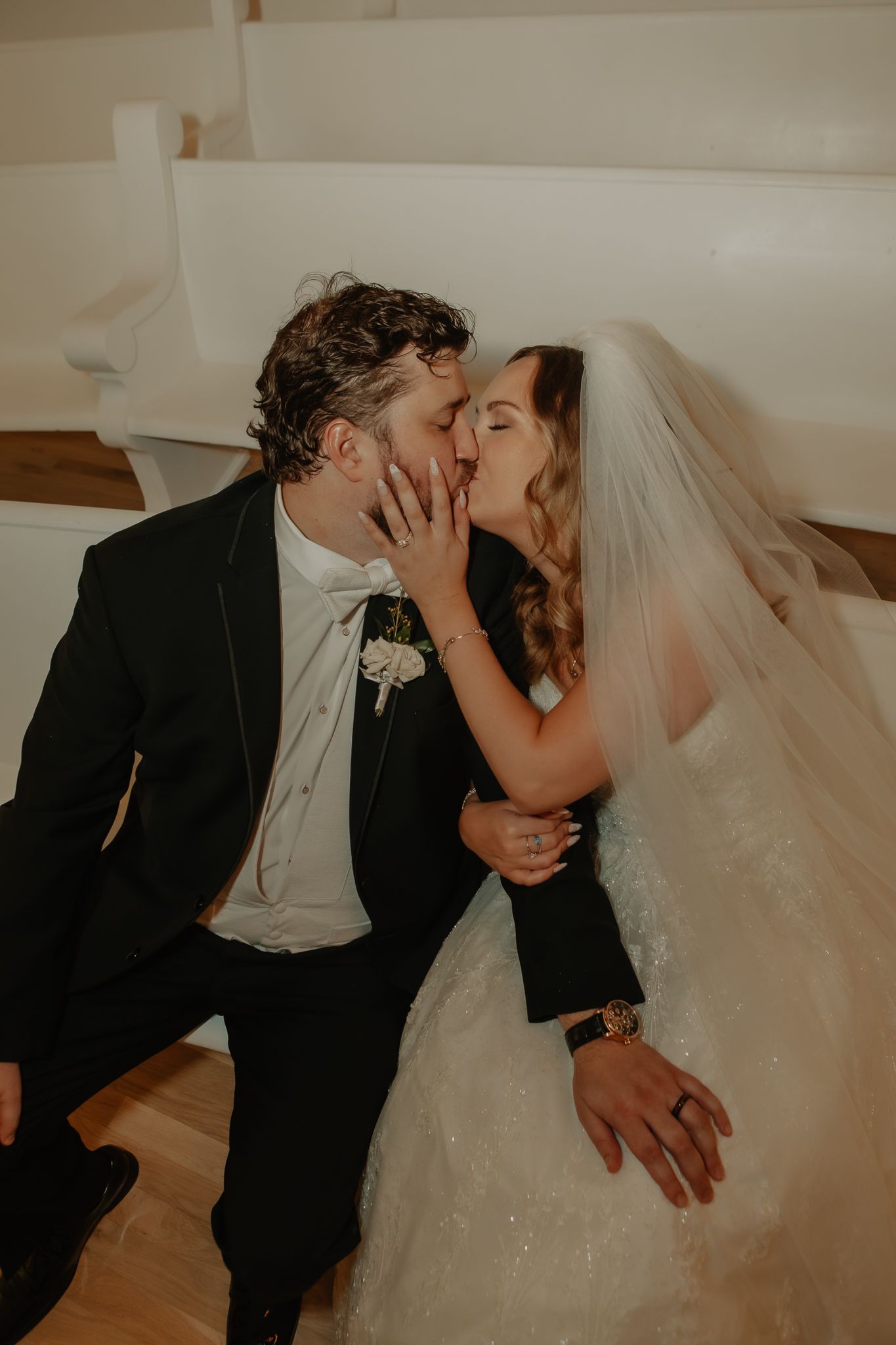 Bride and groom share a romantic kiss in a white chapel, dressed in wedding gown and black tuxedo.
