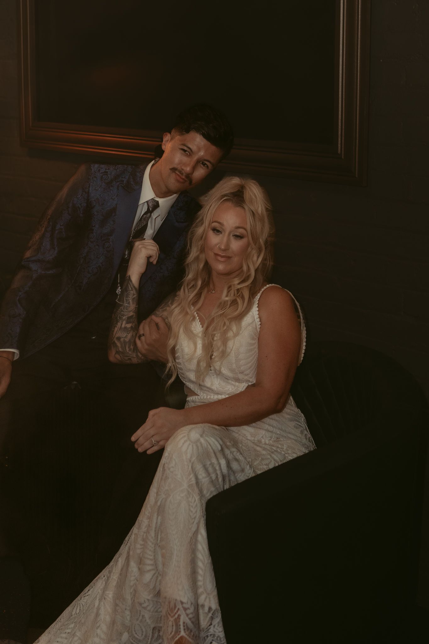 Blonde bride in lace gown leans against tattooed groom in blue floral suit in dramatic dark setting.