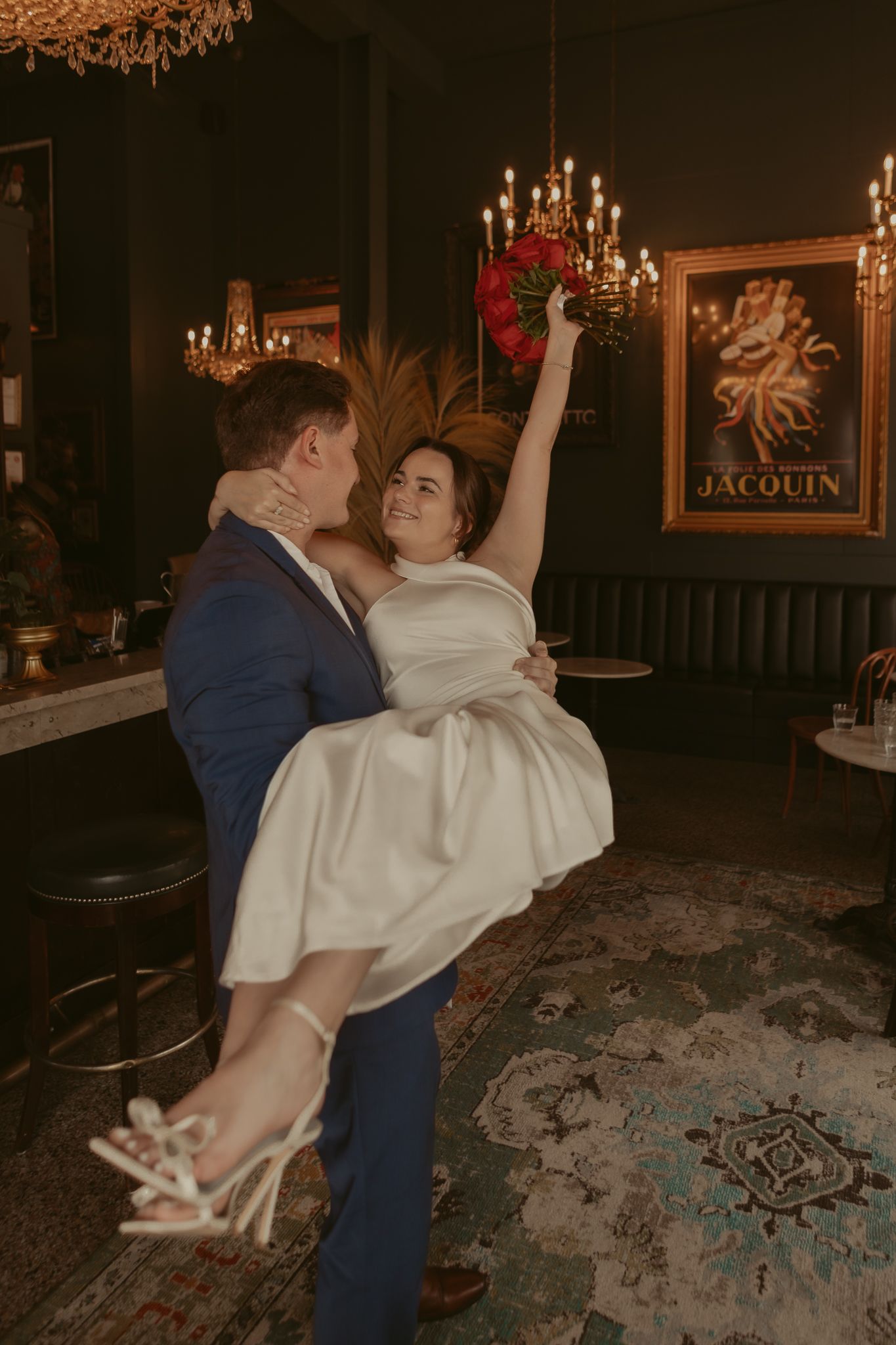 Groom in blue suit lifts smiling bride in white dress holding red bouquet in moody bar.