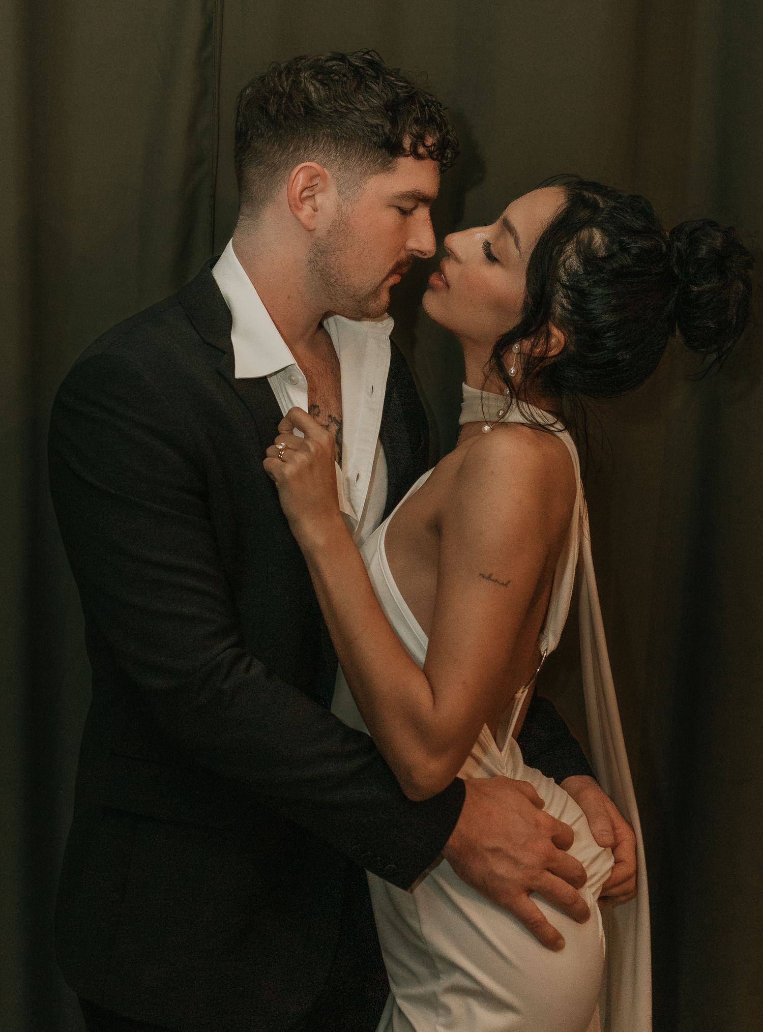 Couple sharing an intimate kiss in formal wedding attire against a dark draped background.