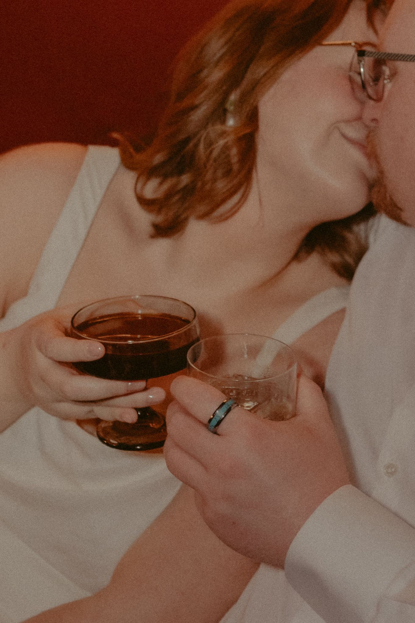 Newlyweds share a kiss while clinking cocktail glasses, bride holding dark drink with wedding ring visible.