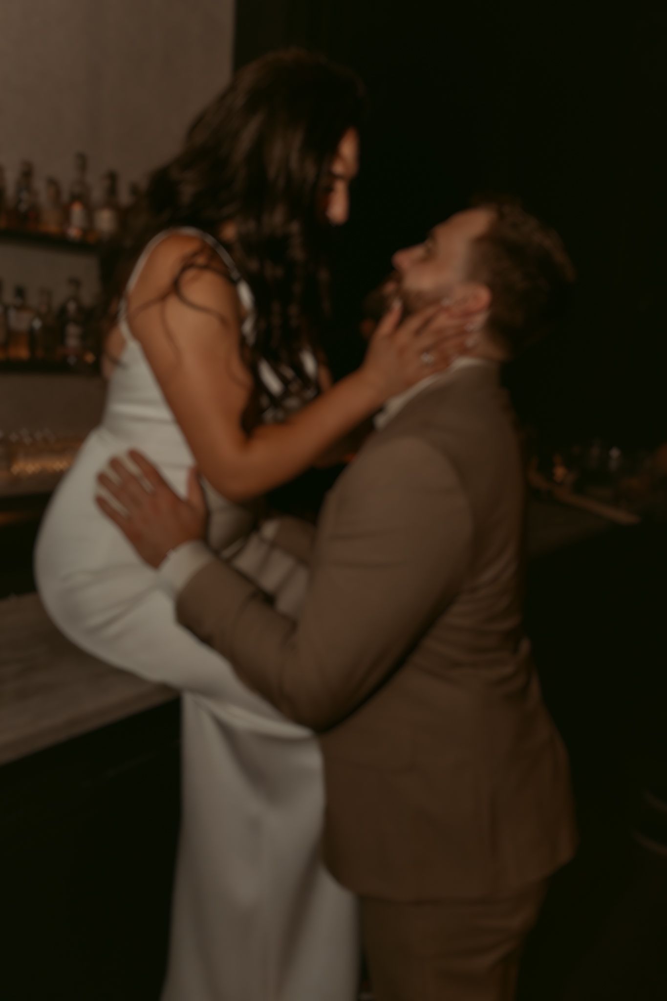 Groom lifts bride in white gown near a bar, sharing a joyful romantic moment at their reception.