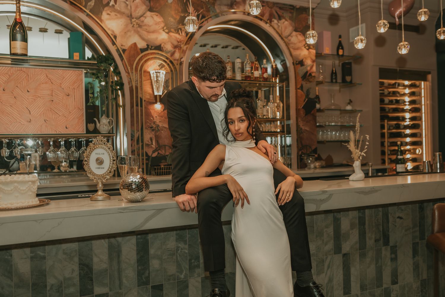 Elegant couple posing at a stylish bar, woman in white gown seated on counter with man in black suit.