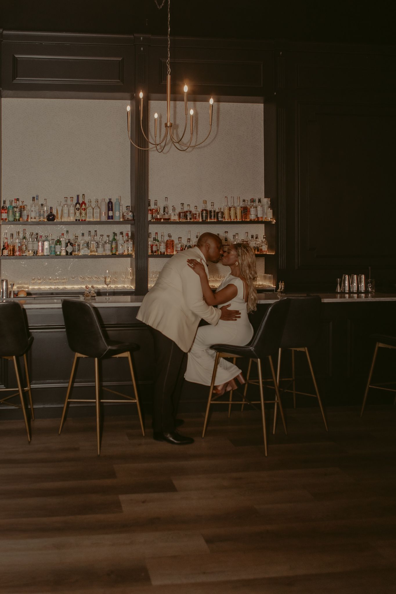 Couple sharing a romantic kiss at an elegant dark bar with candlelight chandelier and shelves of liquor bottles.