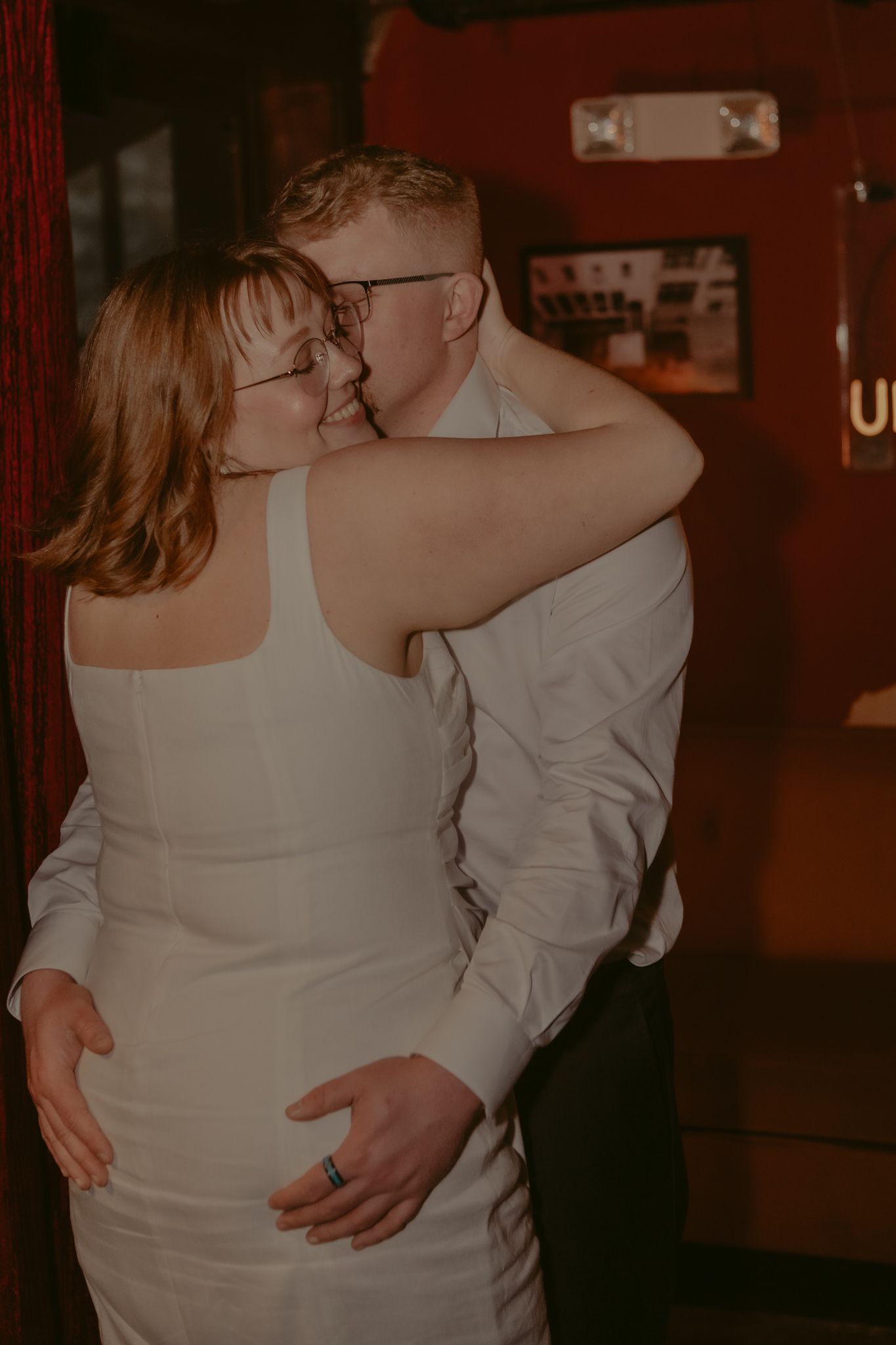 Groom kisses bride tenderly as she wraps arms around him in warmly lit wedding reception setting.