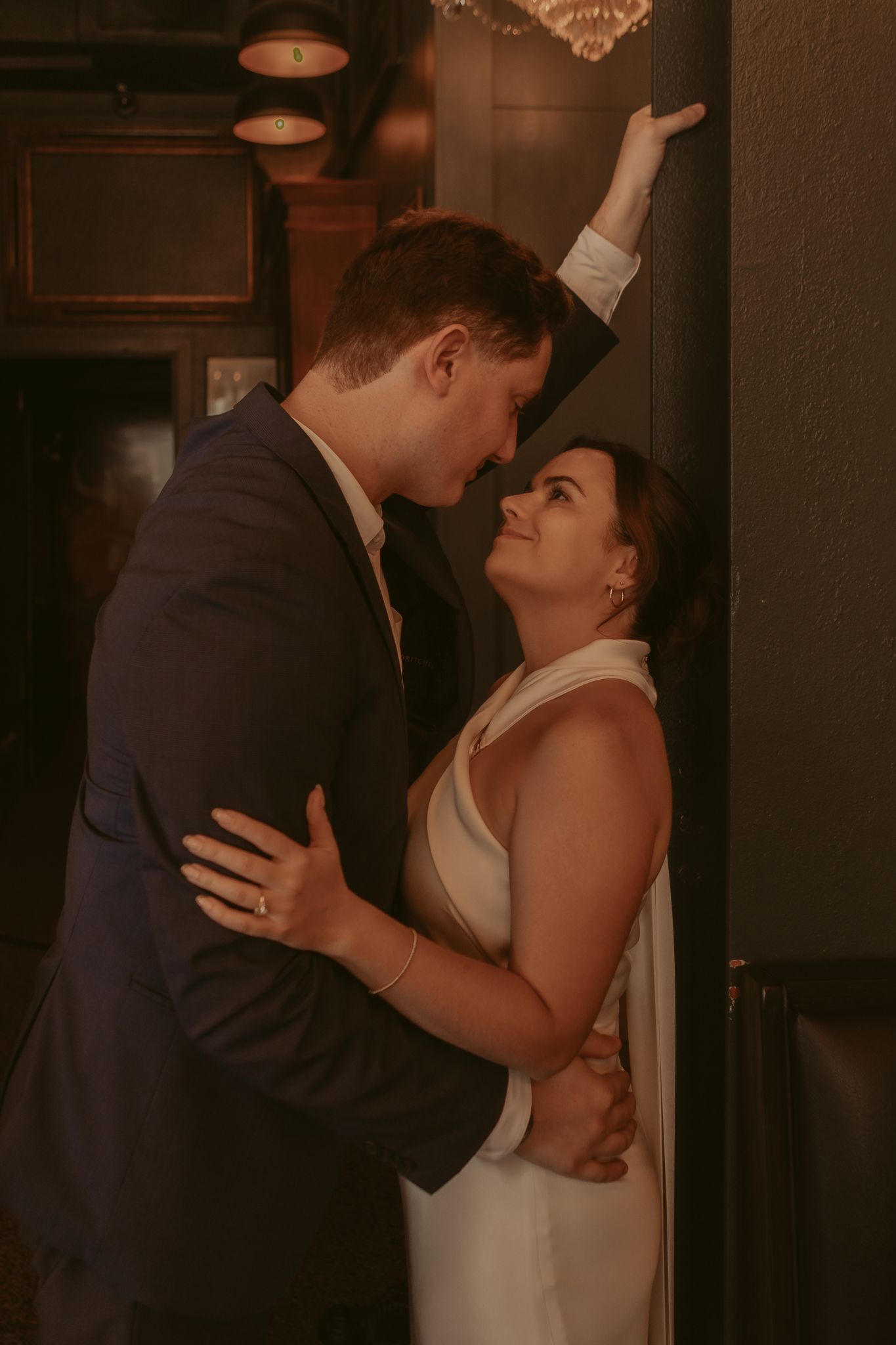 Groom in navy suit leans toward smiling bride against dark wall in intimate bar setting.