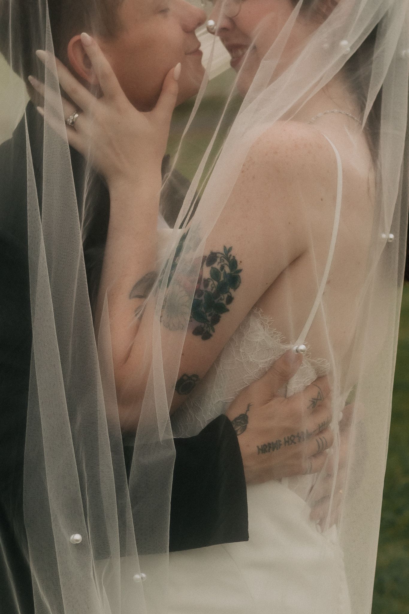 Bride and groom share intimate kiss under sheer wedding veil, bride's tattooed arms visible in romantic close-up.