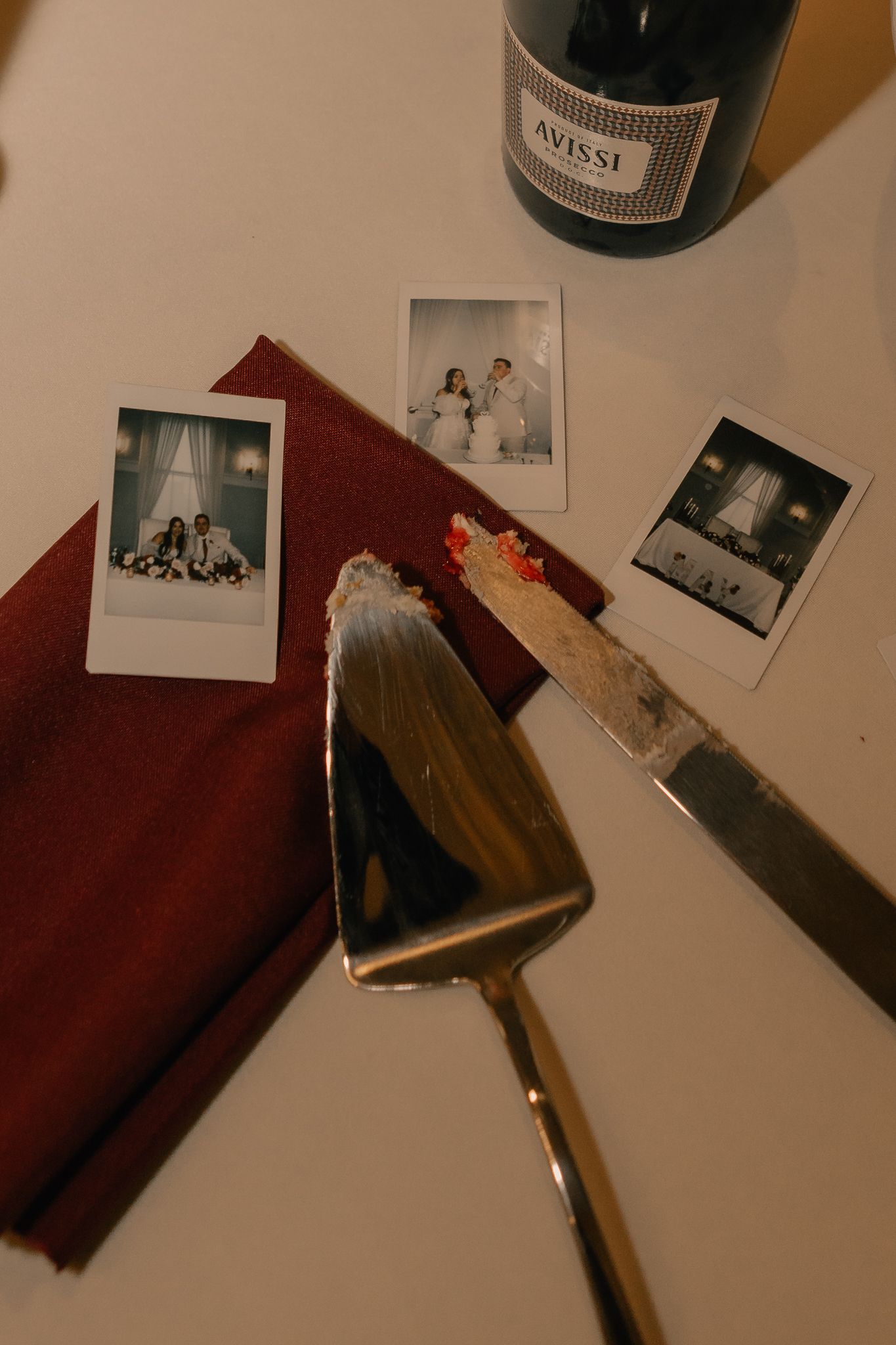 Wedding cake cutting utensils beside Polaroid photos and Avissi champagne bottle on white table.