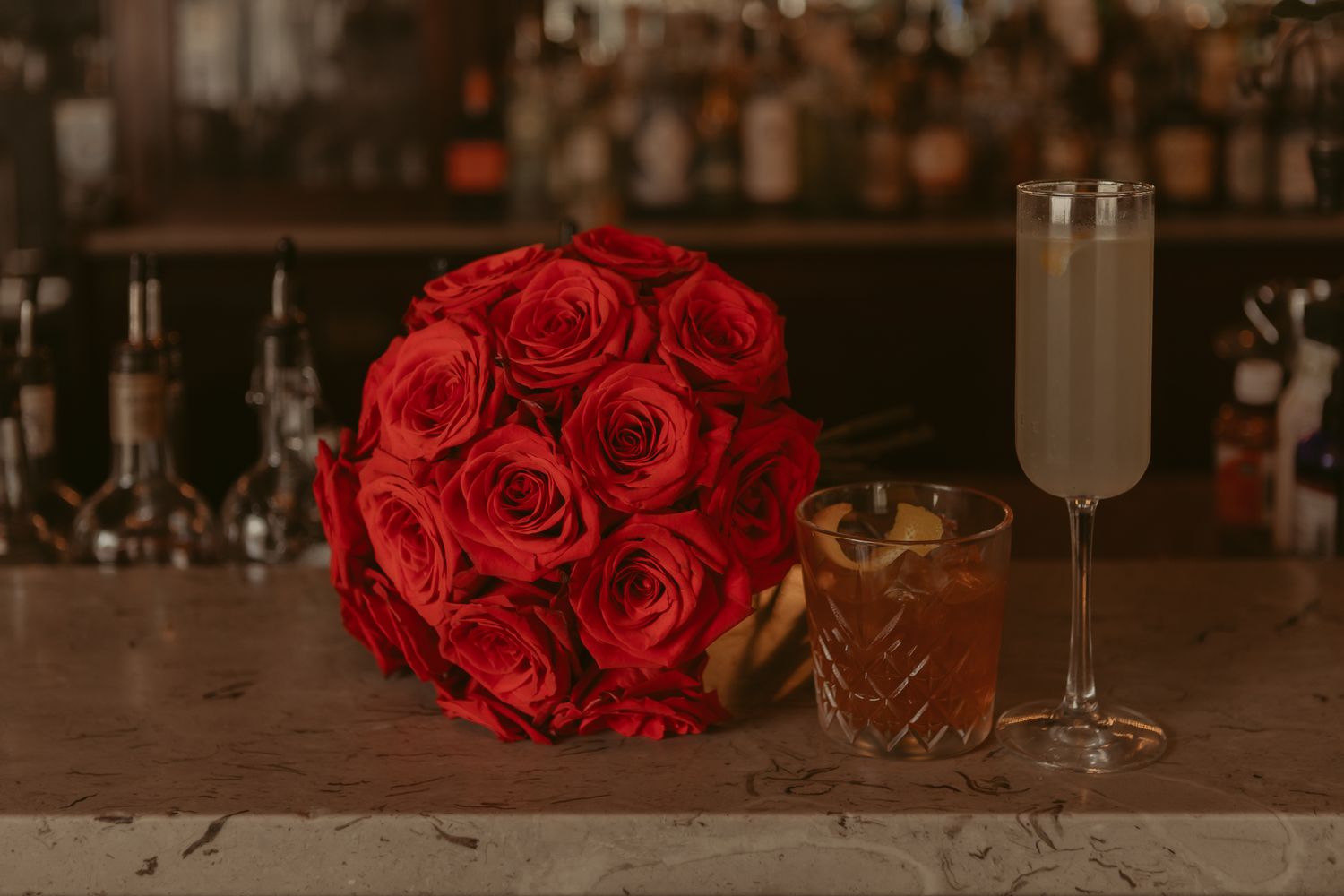 Lush red rose bridal bouquet beside champagne flute and whiskey cocktail on marble bar.