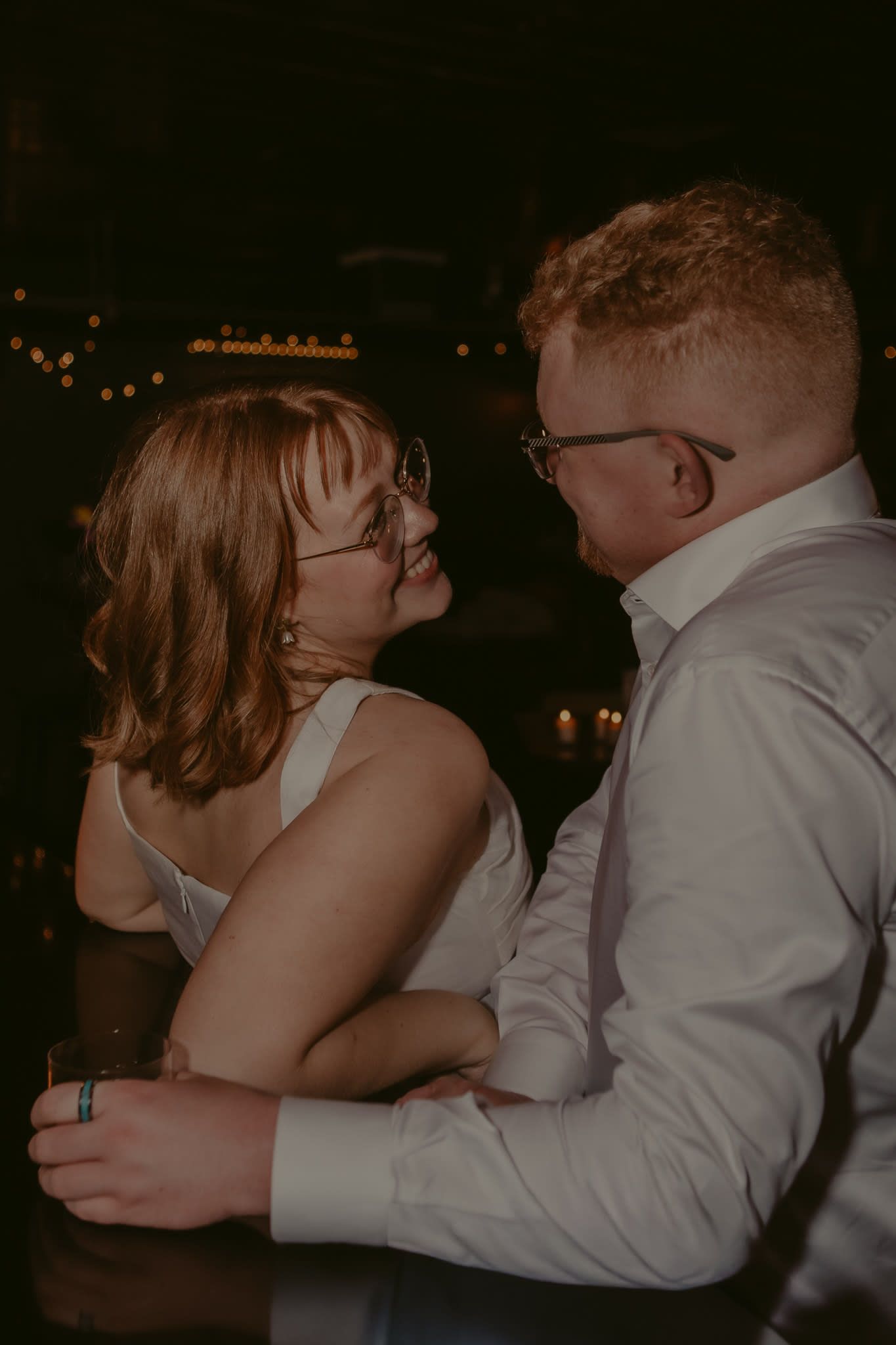Couple slow dancing together at a dimly lit wedding reception, smiling at each other while wearing glasses.