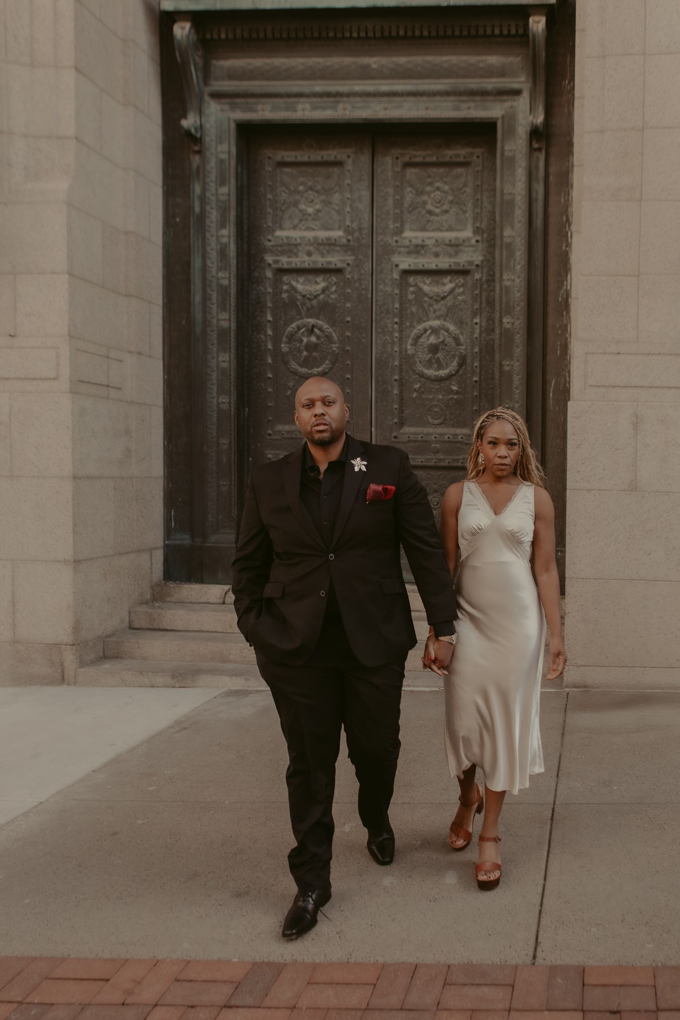 Couple in formal attire walking away from ornate bronze doors, man in black suit, woman in ivory slip dress.
