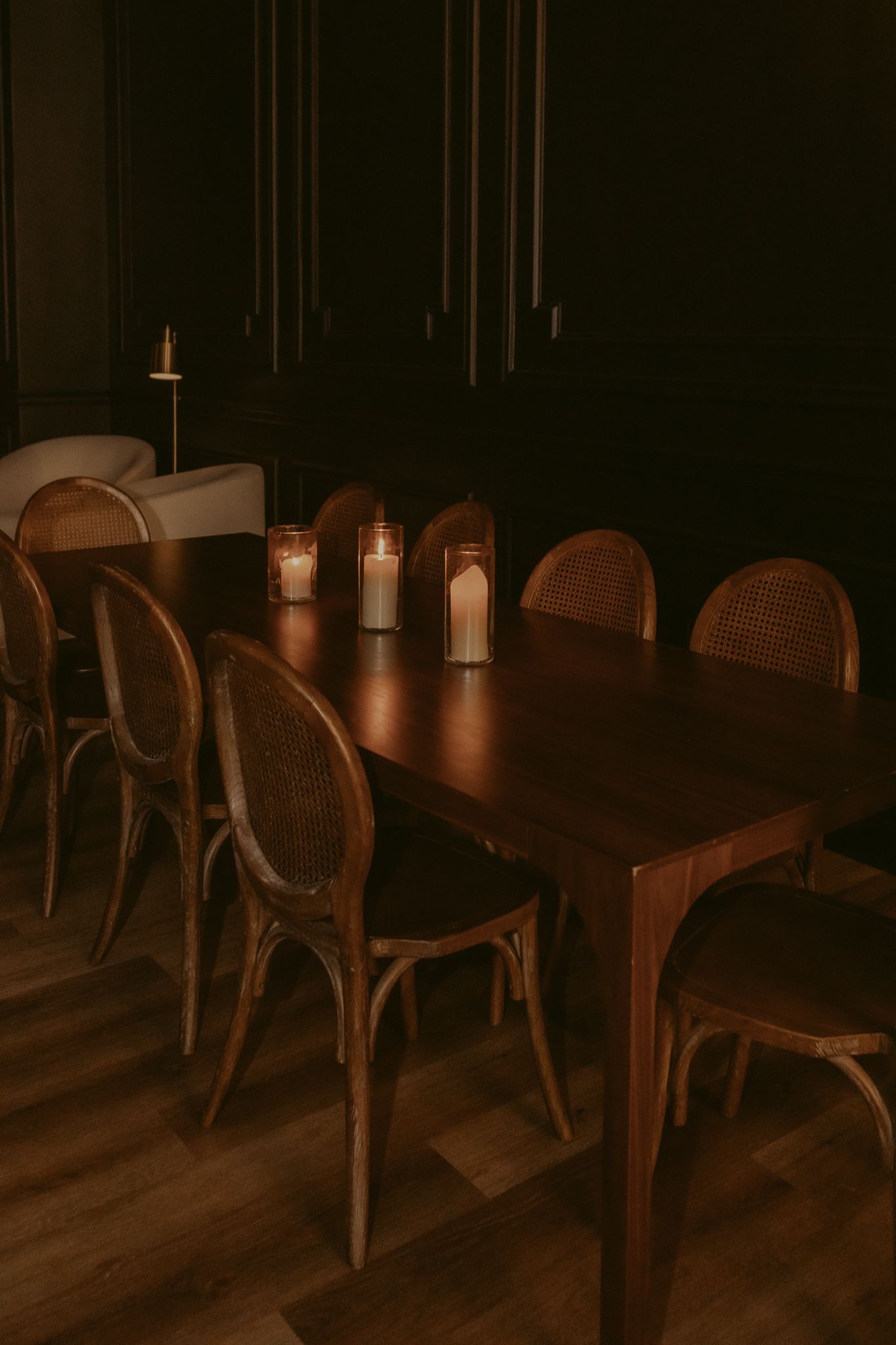 Dark, moody private dining room with wooden table, rattan chairs, and glowing candle centerpieces.