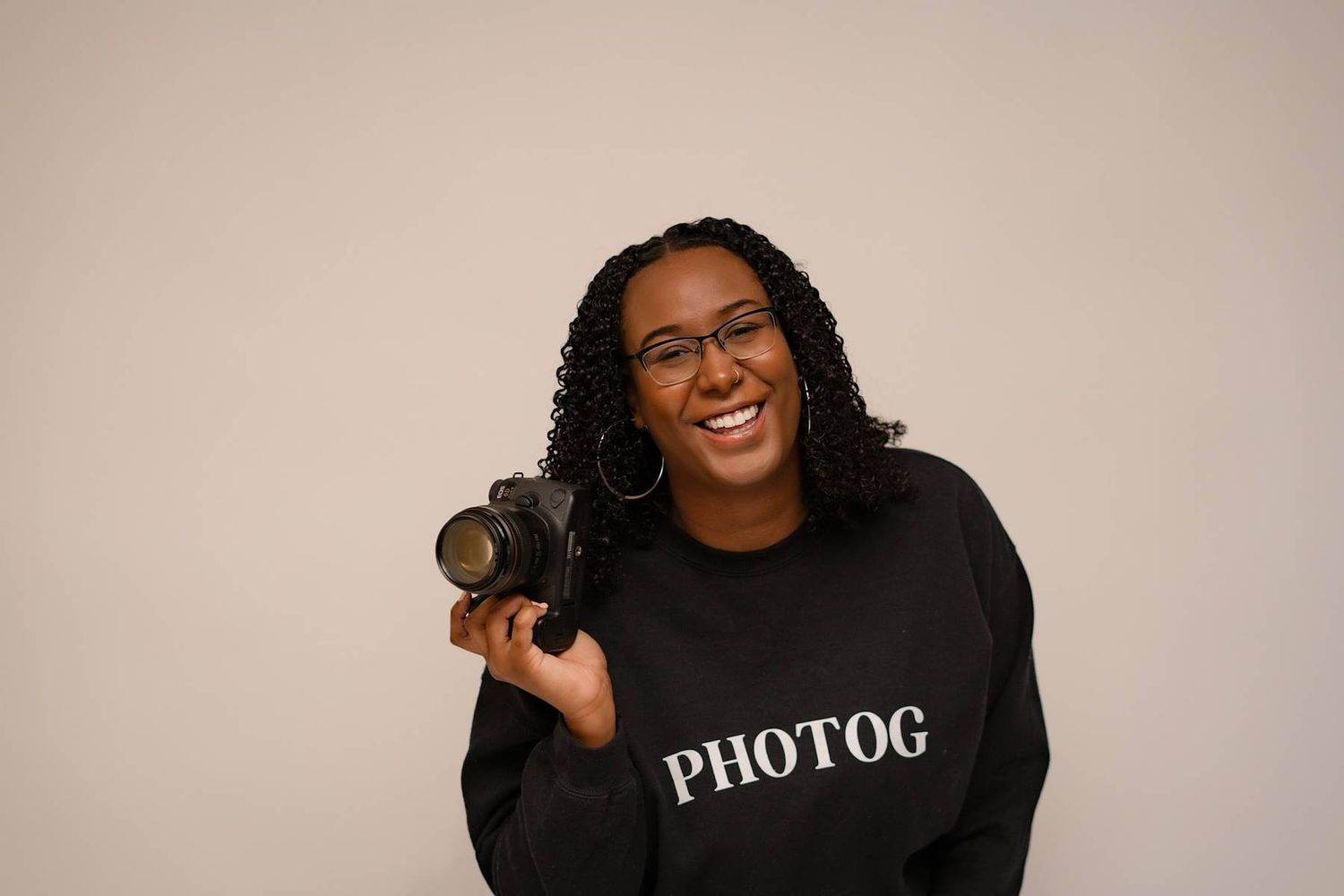 Smiling female photographer in PHOTOG sweatshirt holding DSLR camera against neutral background.