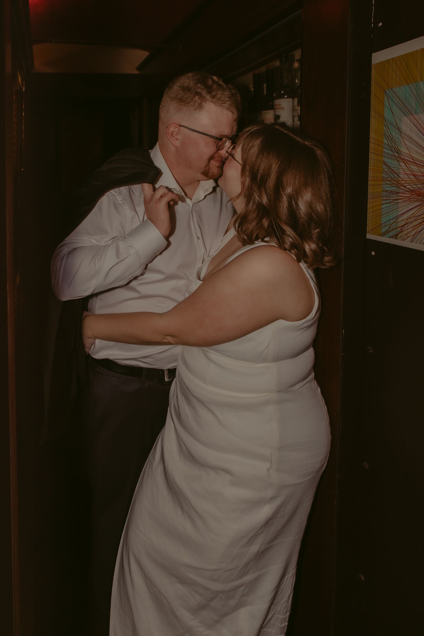 Bride and groom share a passionate kiss in dimly lit venue, bride in sleeveless white gown.
