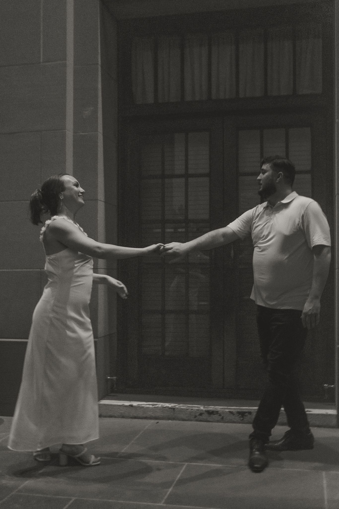 Bride and groom hold hands dancing outdoors in black and white wedding photo near building entrance.