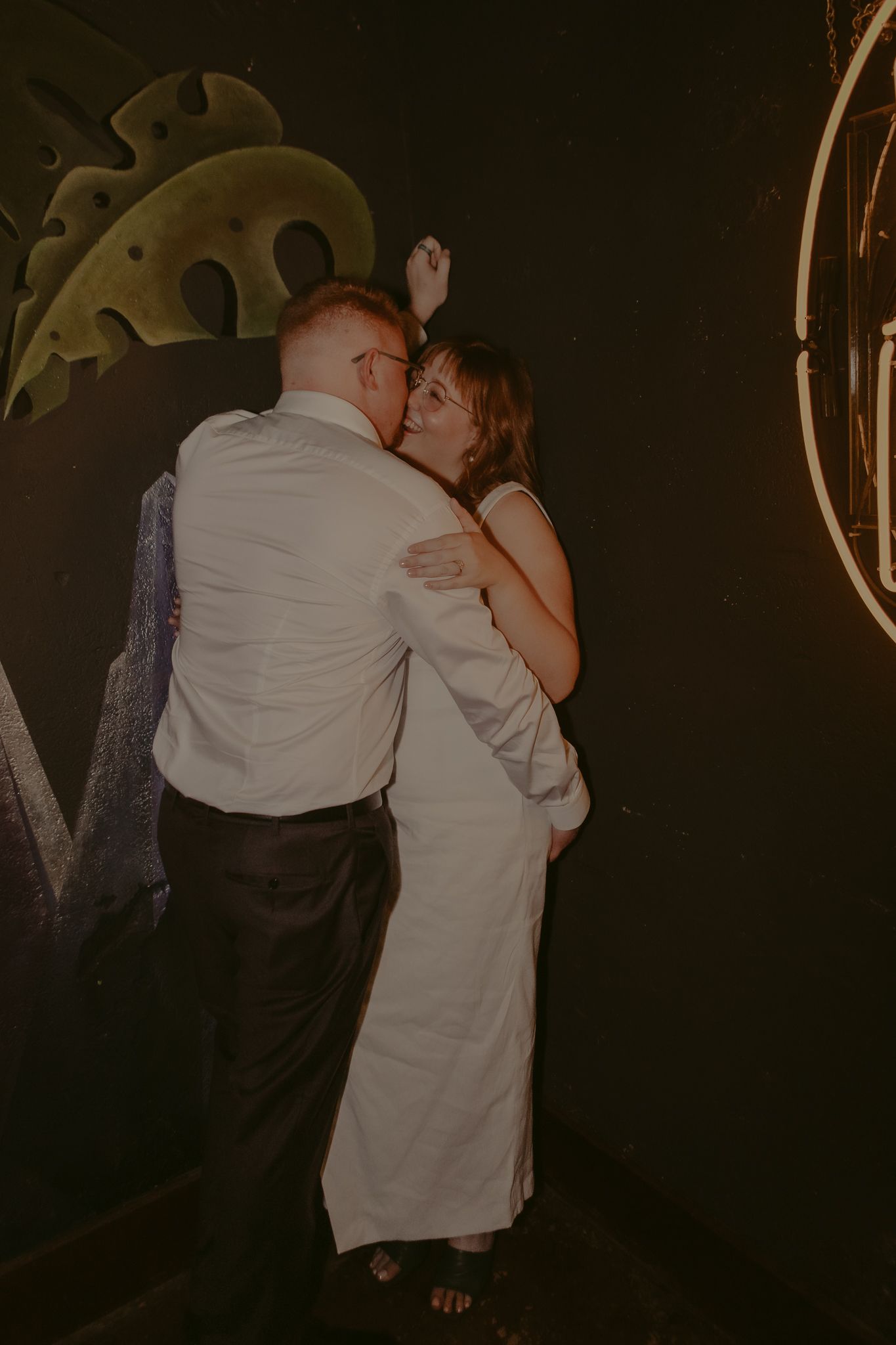 Newlyweds embrace and dance together against dark mural wall in moody, warmly lit reception venue.