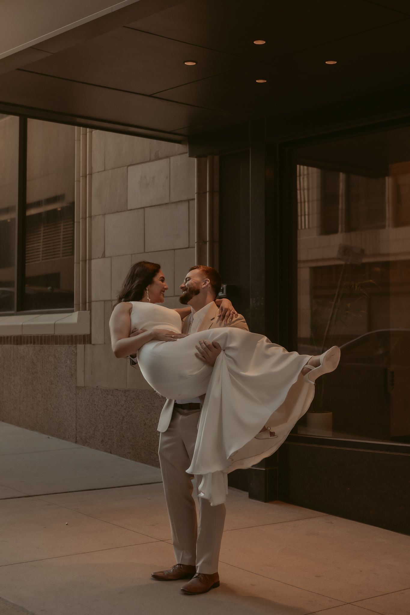 Groom lifts bride in white wedding dress outside urban building at golden hour.