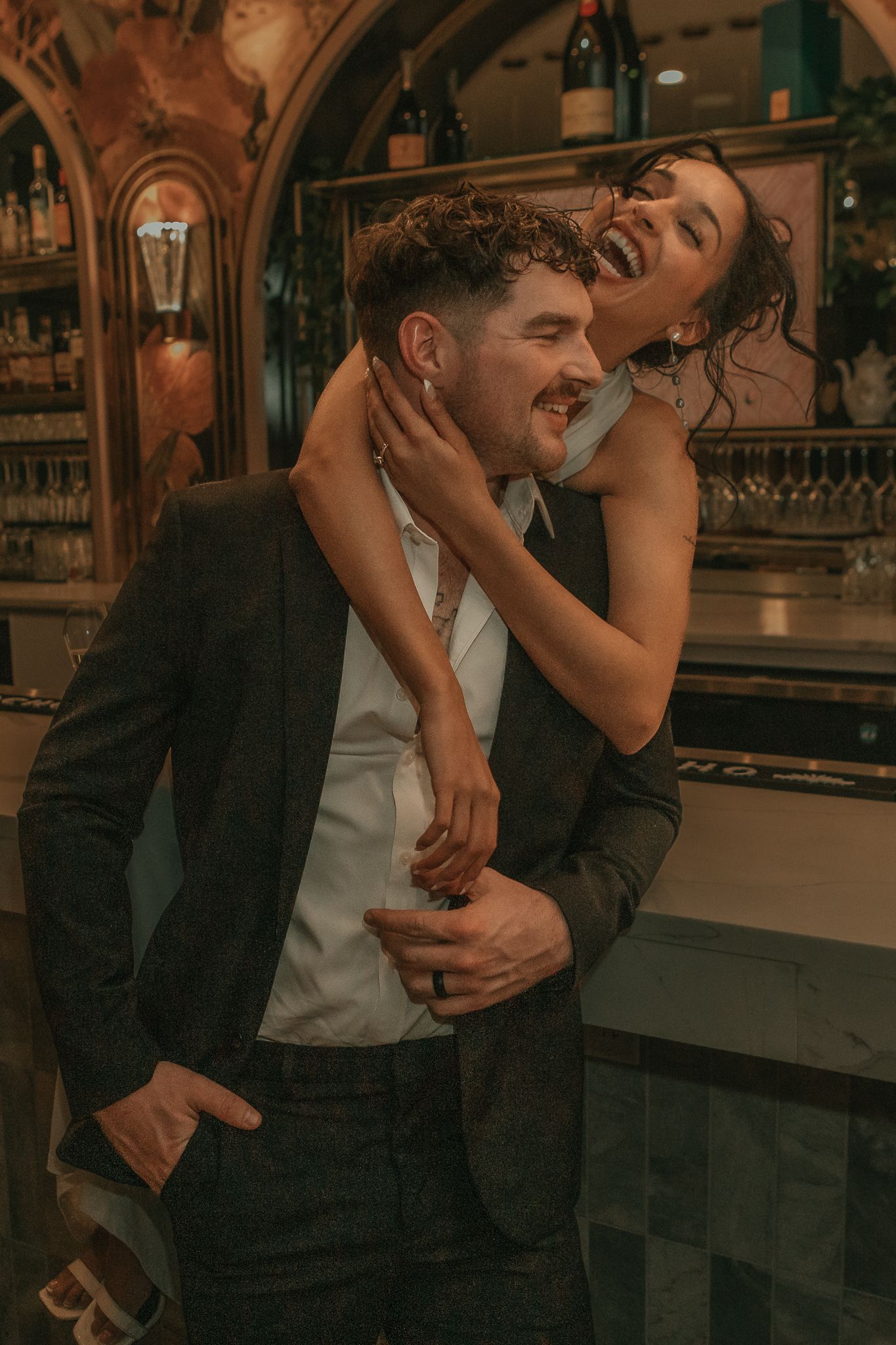 A couple laughing and embracing romantically at an elegant bar, man in suit, woman in black dress.