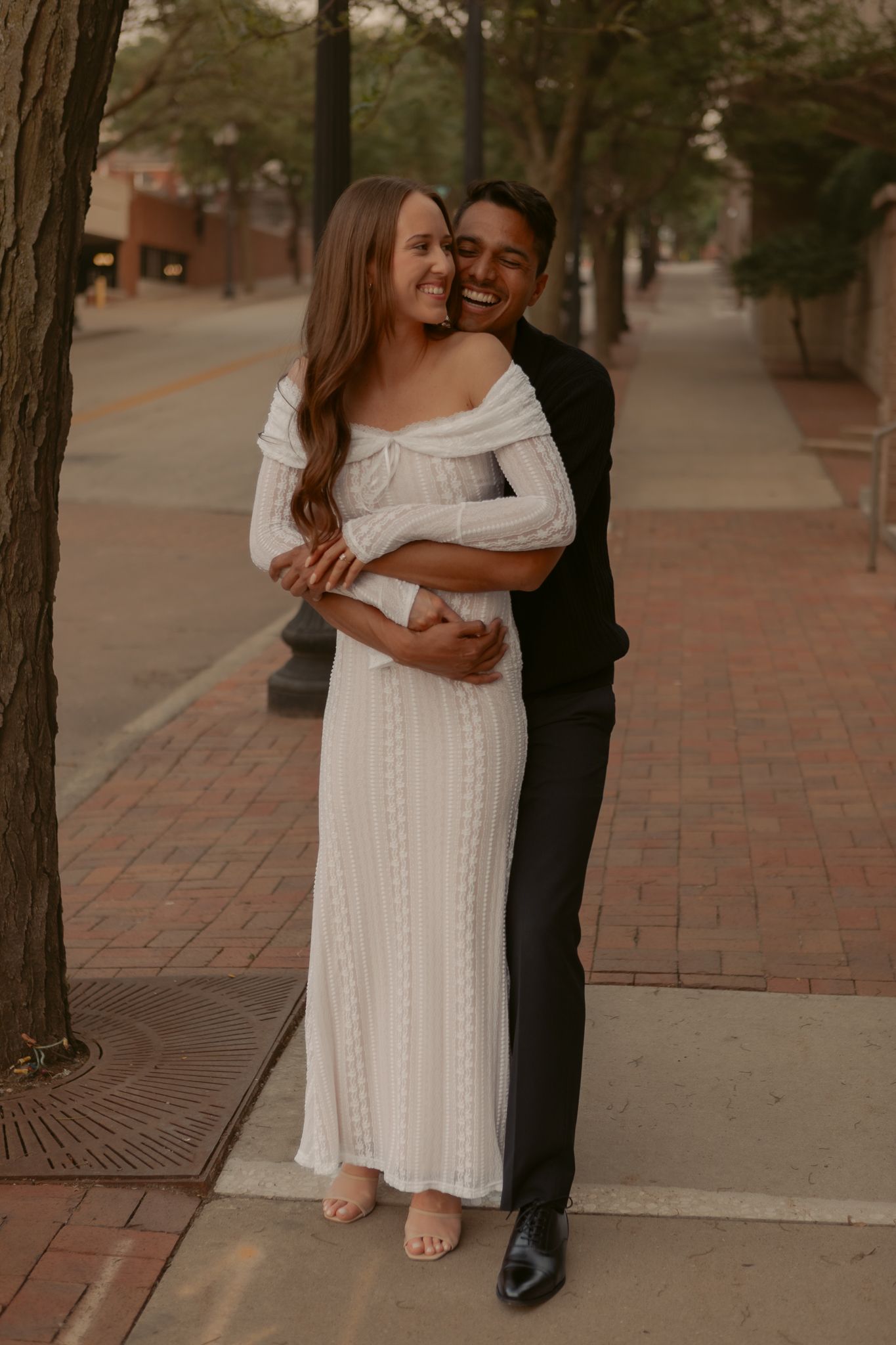 Happy couple embracing on a brick sidewalk, woman in white off-shoulder dress and man in black suit smiling.