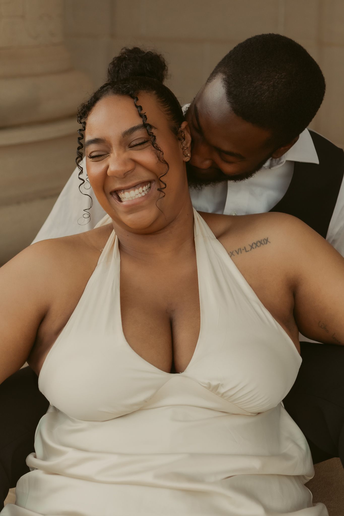 Joyful bride in white halter gown laughing as groom kisses her neck during wedding portraits.
