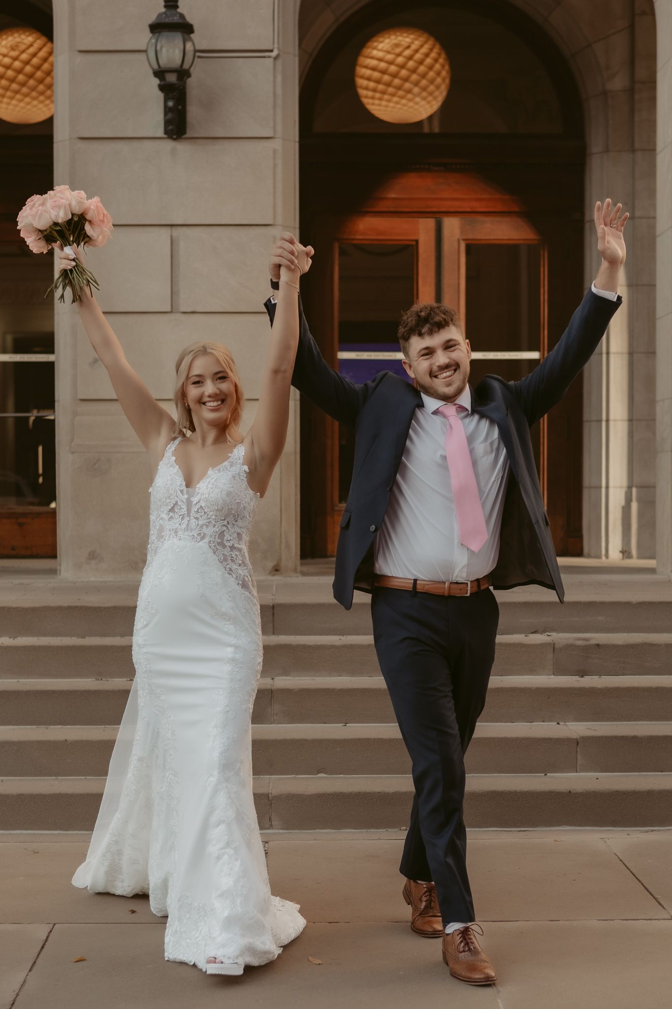 Joyful newlyweds raising hands on building steps, bride in lace gown holding pink bouquet, groom in navy suit.