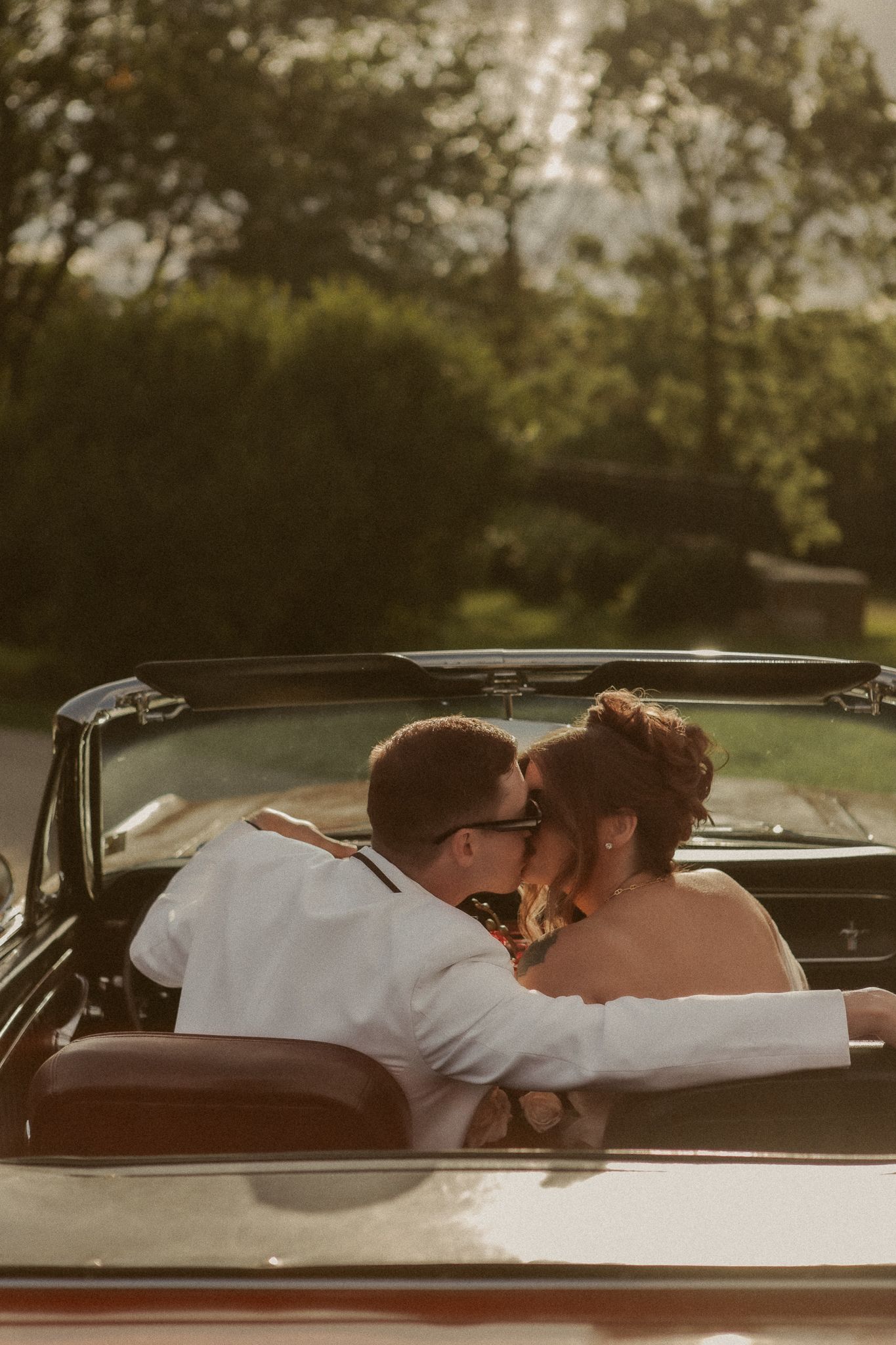 Newlyweds share a romantic kiss in a classic convertible car at golden hour sunset.