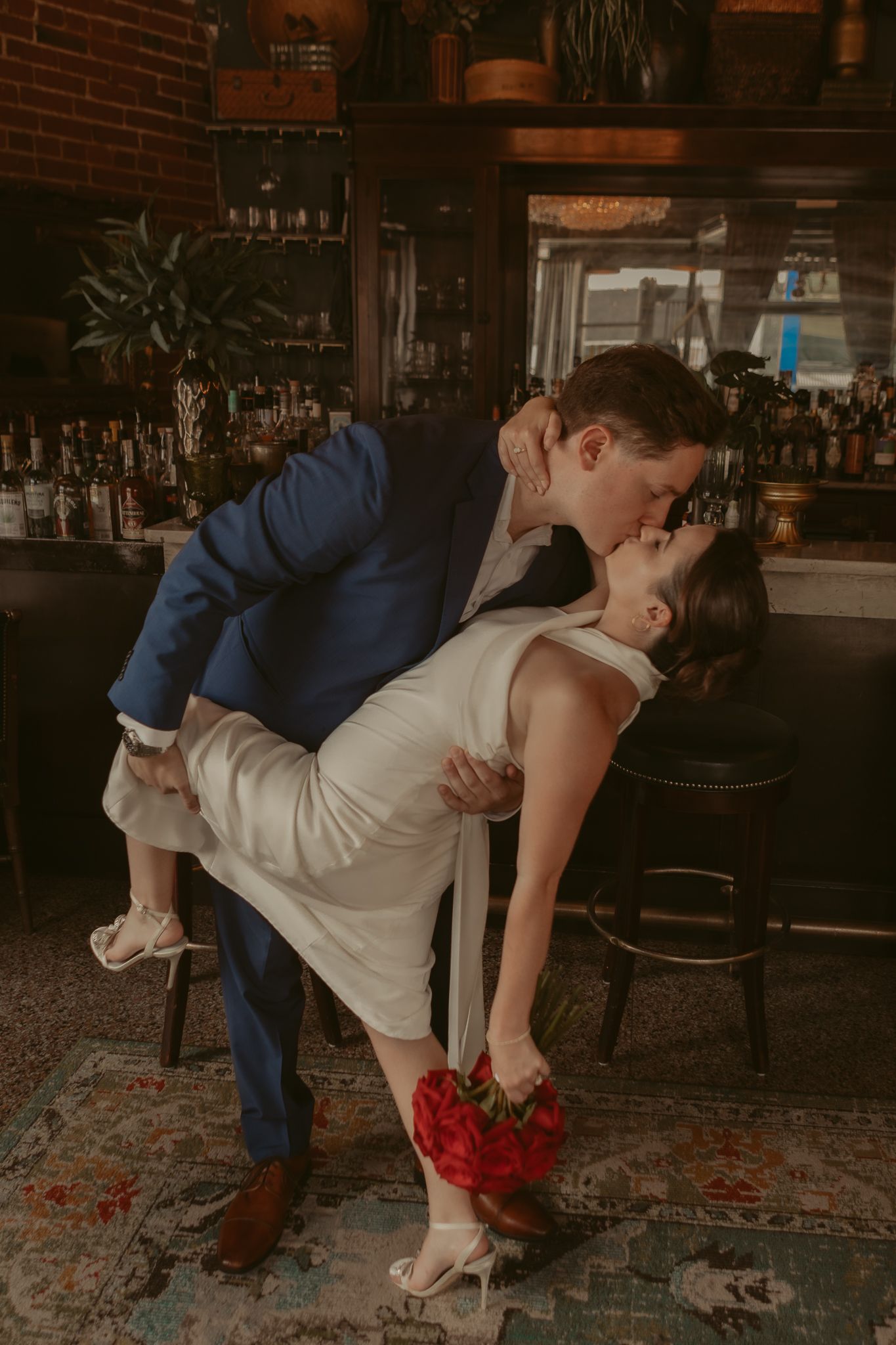 Groom in blue suit dipping bride for a kiss at a bar, bride holding red bouquet.