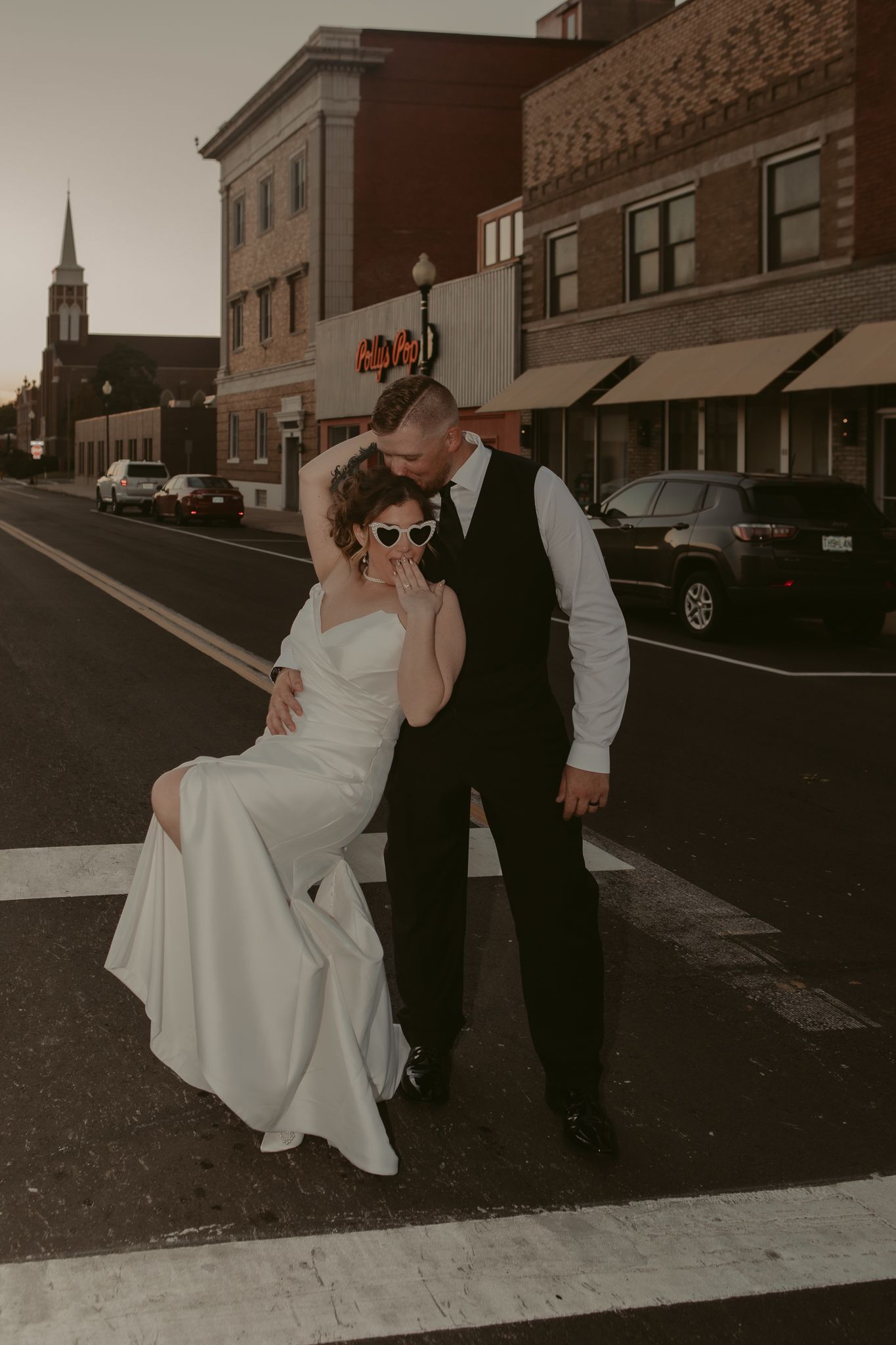 Bride in white satin gown with sunglasses and groom in black vest embrace on a crosswalk at sunset.