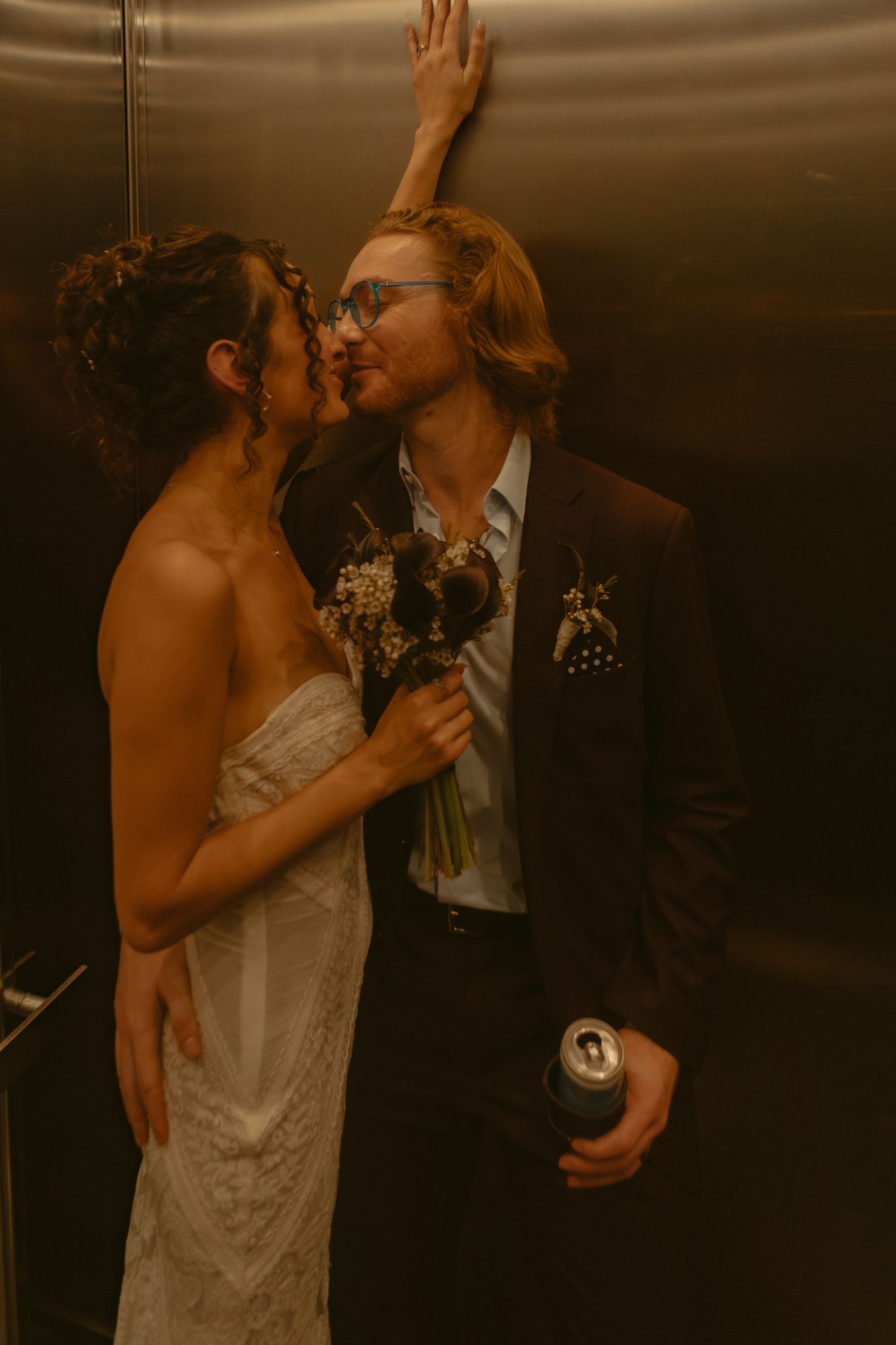 Bride and groom share a romantic kiss in a golden elevator, groom holding a bouquet and a can.