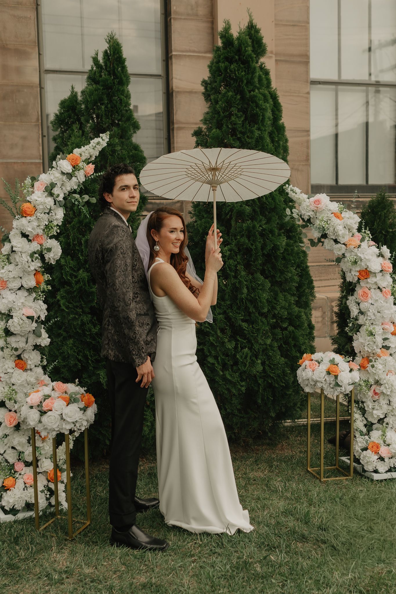 Bride in white gown holds parasol with groom before floral arch with roses outdoors.