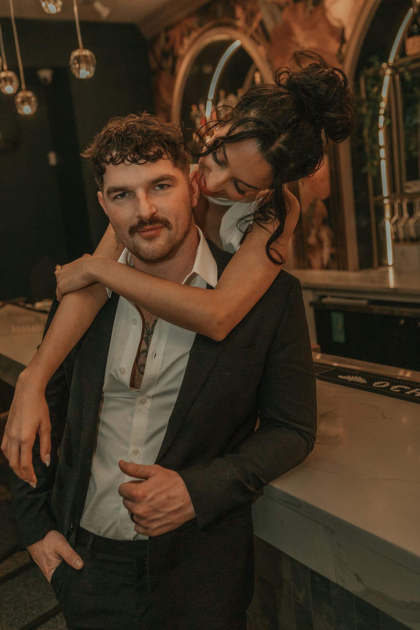 Bride drapes arms over groom shoulders from behind in dark elegant bar interior setting.