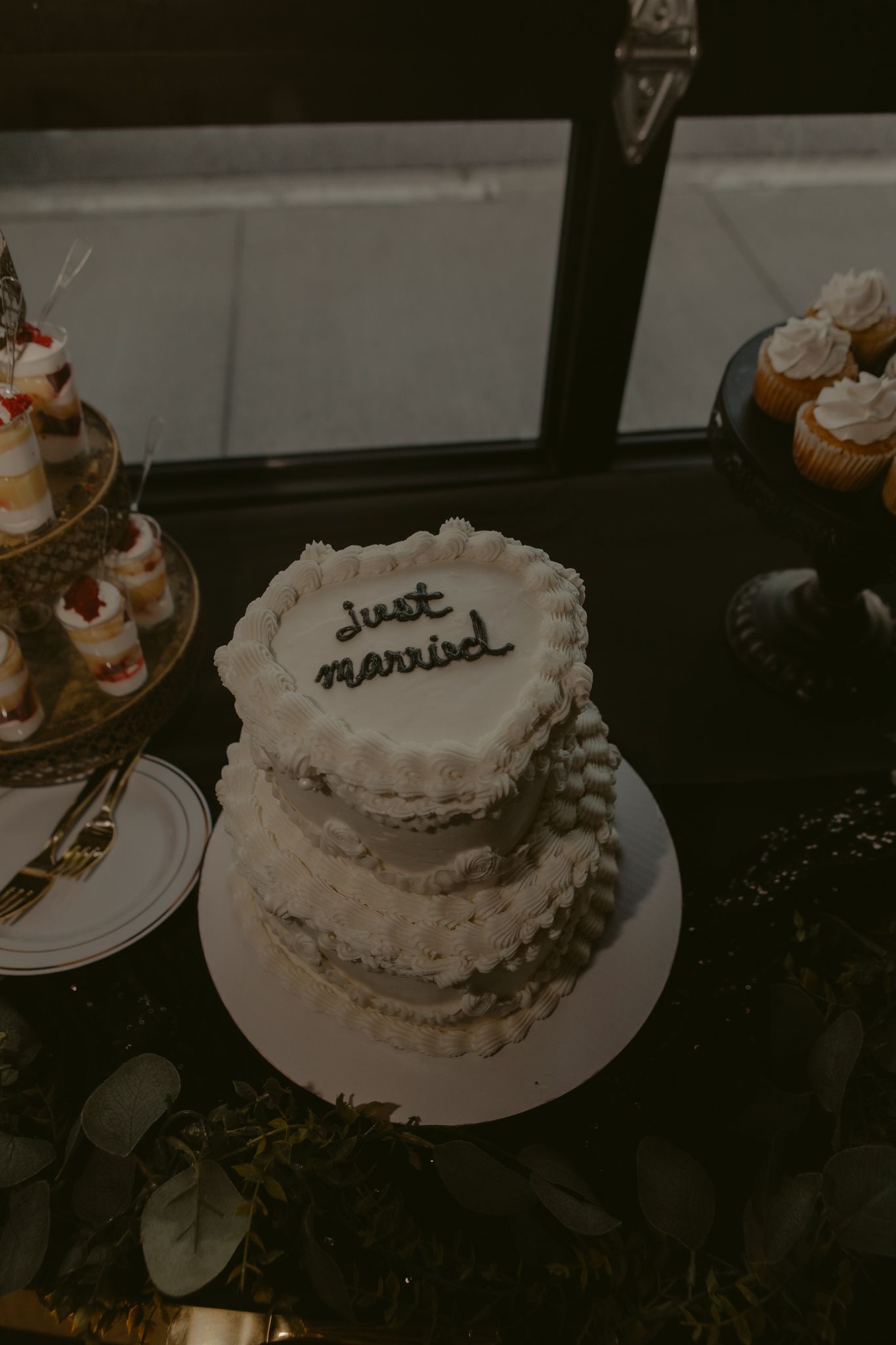 Two-tier heart-shaped white wedding cake with just married inscription surrounded by desserts.