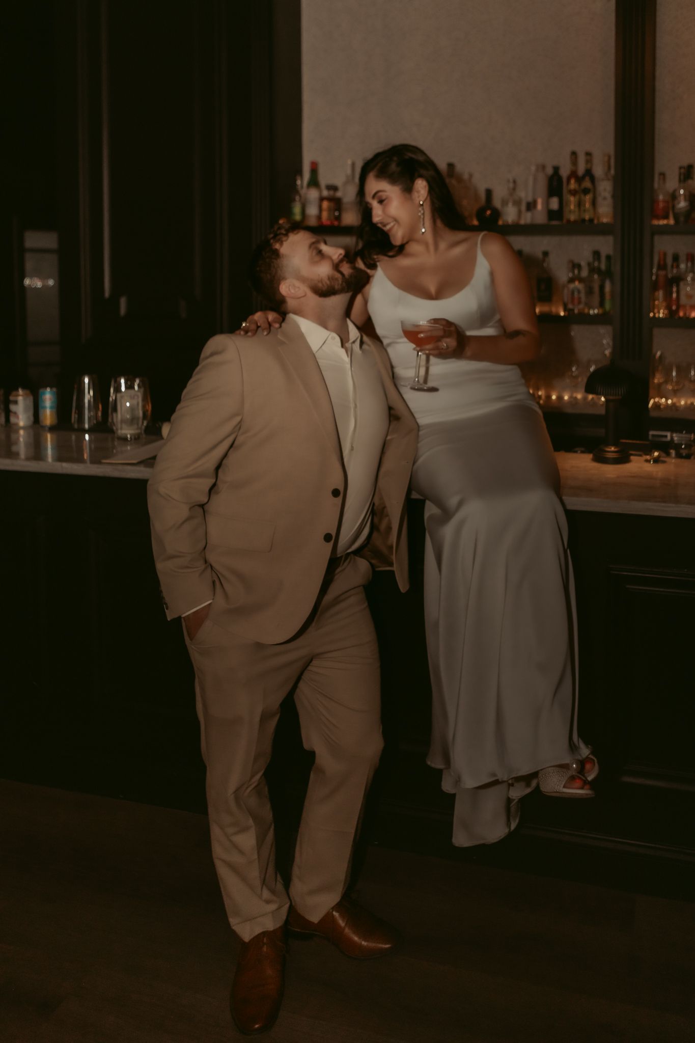 Bride and groom share a playful moment at a dimly lit bar, she in a white dress, he in a tan suit.