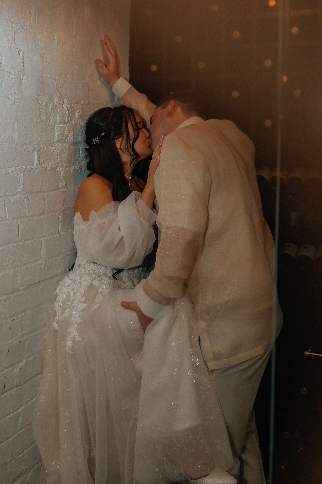 Groom in linen suit leans in to kiss bride in off-shoulder floral gown against white brick wall.