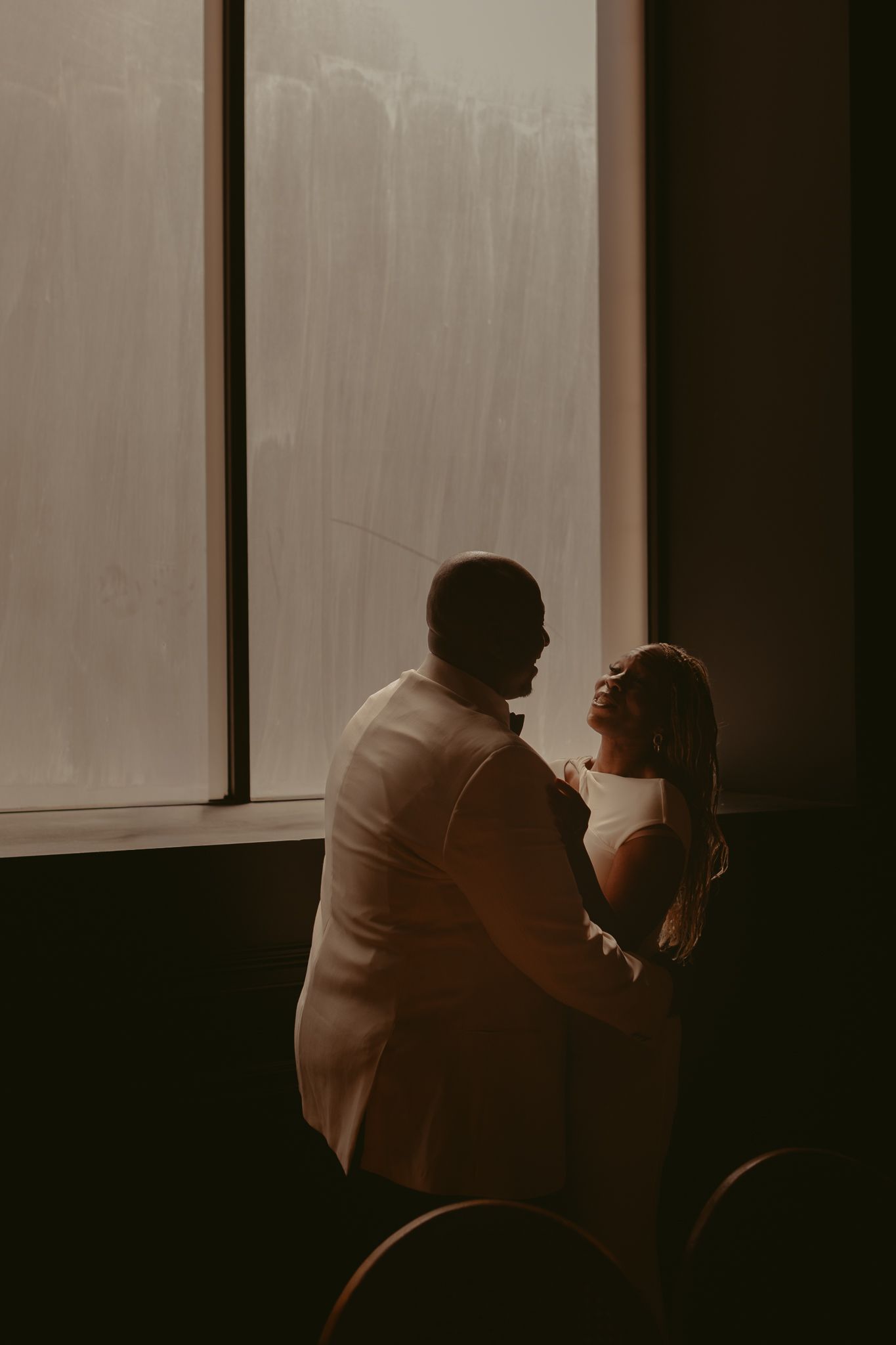 Silhouette of bride and groom sharing romantic dance in front of large sunlit window.