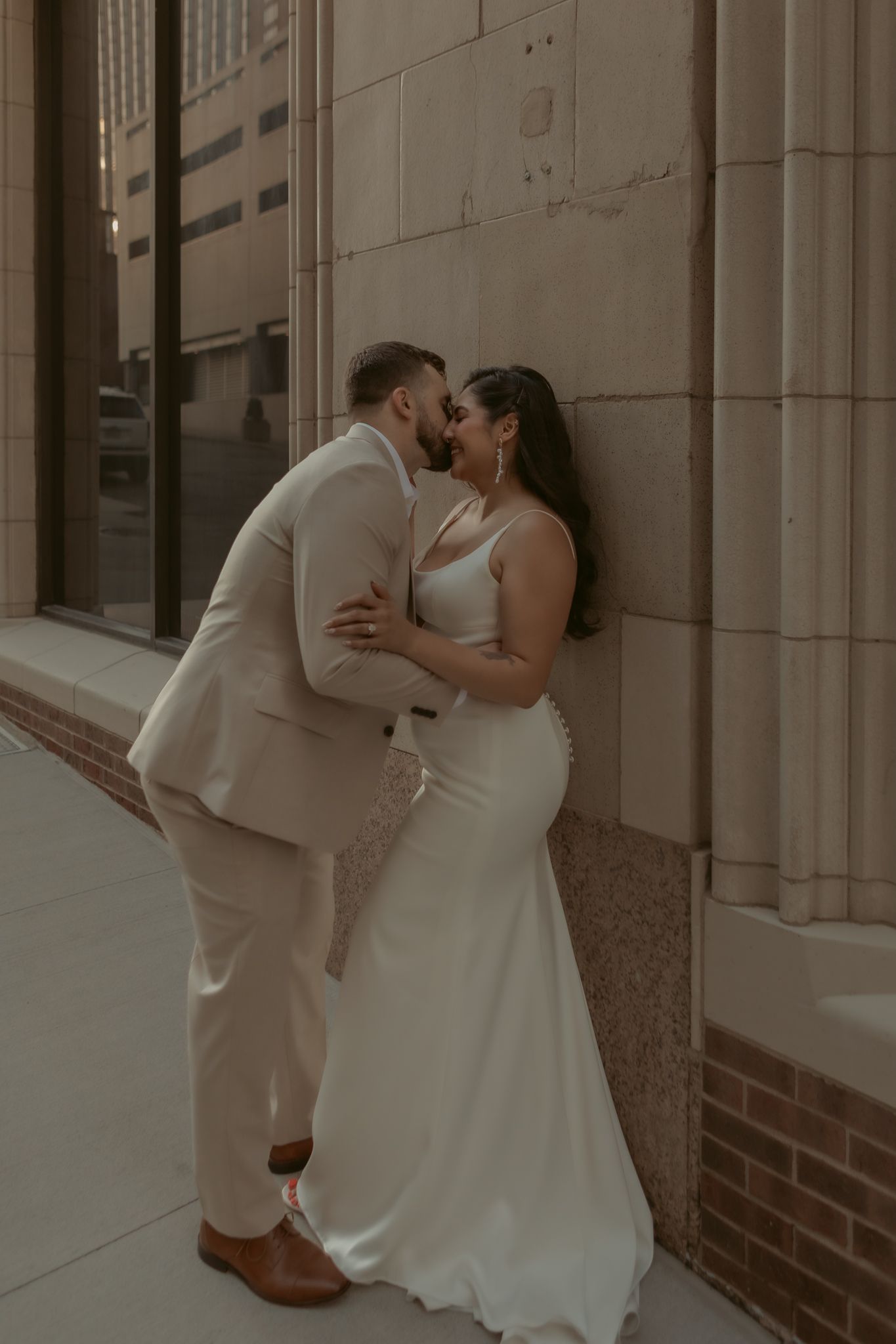 Bride and groom sharing a kiss against a stone building, groom in beige suit and bride in fitted white gown.
